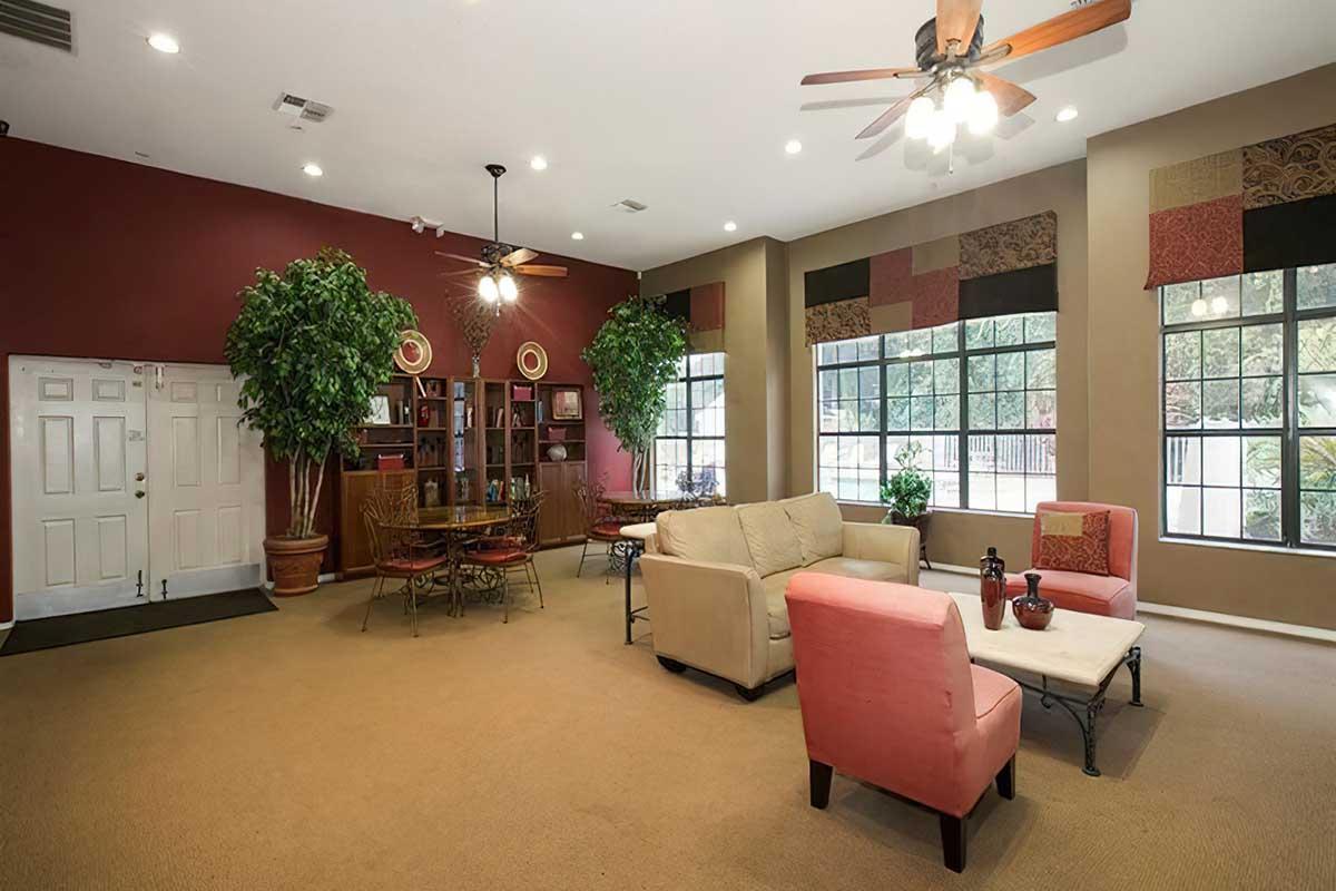 A spacious lounge area featuring two ceiling fans, large windows with multi-colored valances, potted plants, and a mix of seating arrangements including a beige sofa, a pink armchair, and a coffee table. The decor is warm and inviting, with a cozy feel highlighted by soft lighting.