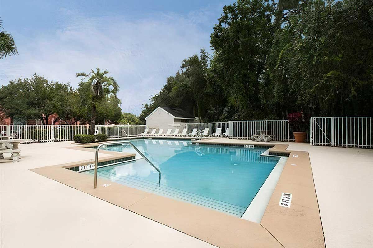 A clear swimming pool surrounded by a fenced area, with lounge chairs arranged along the poolside. Lush green trees and foliage provide a scenic backdrop, creating a relaxing outdoor atmosphere. The pool features a railing and is well-maintained, inviting visitors to enjoy a refreshing swim.