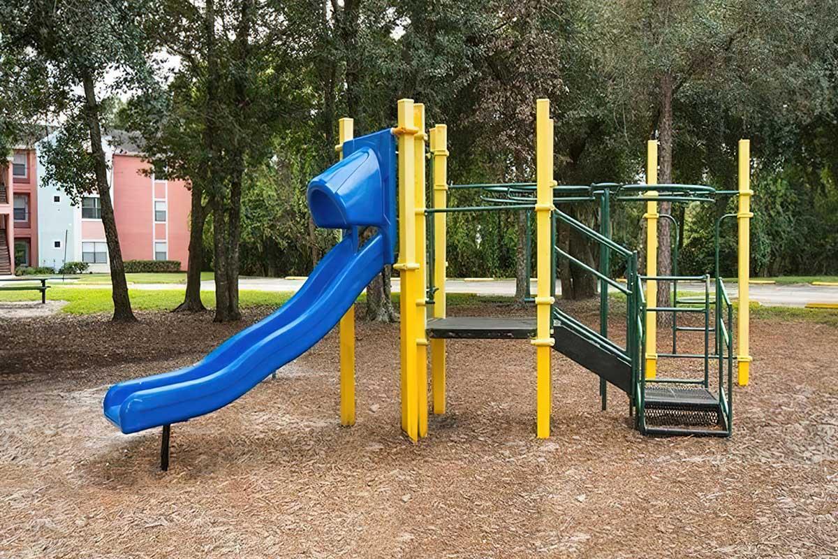 A blue playground slide attached to a green and yellow climbing structure, surrounded by trees and grassy areas. The scene is set in a park with playground equipment designed for children to play and explore.