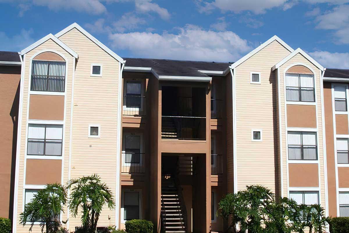 A view of a three-story apartment building with beige and light brown siding, featuring balconies and ground-level staircases. Lush green landscaping with palm-like plants surrounds the building under a partly cloudy blue sky.