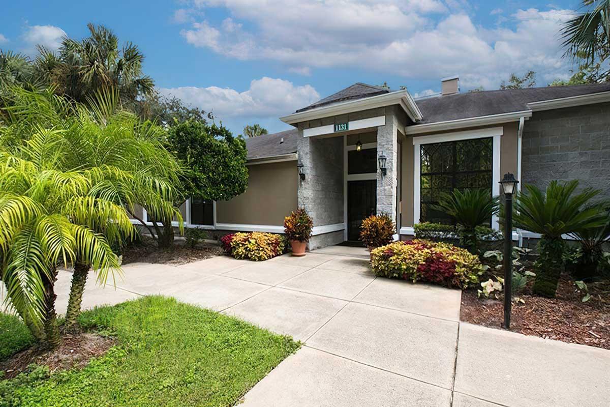 A welcoming modern house with a stone facade and a well-maintained garden. The entrance features lush palm trees and colorful flowering plants along a clean pathway. Bright blue skies and white clouds create an inviting atmosphere.