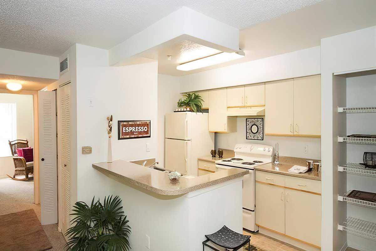 A modern kitchen with white cabinetry and a breakfast bar. It features a stove, an oven, and various kitchen appliances. A small plant decorates the counter, and there's a sign with the word "ESPRESSO." The open layout connects to a living area in the background, enhancing the spacious feel.
