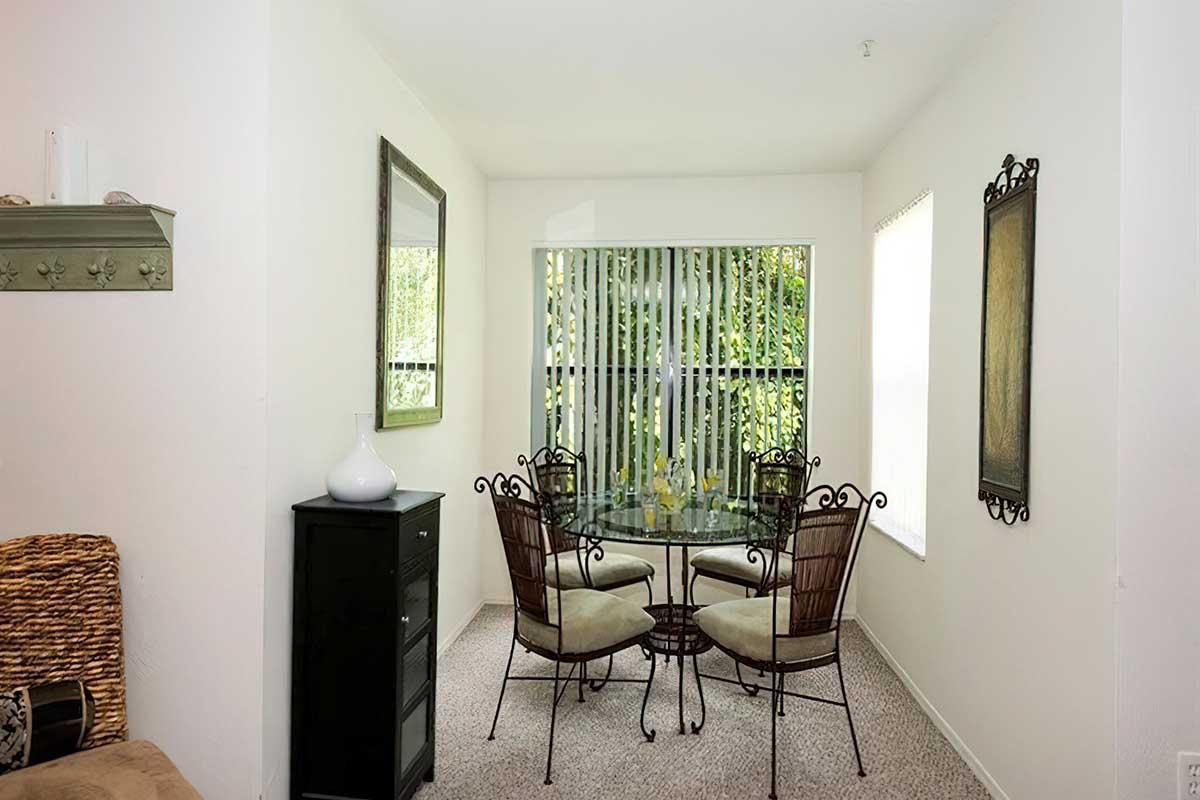 Bright dining area featuring a glass table surrounded by four metal chairs with cushions. A tall cabinet is to the left, and there are large windows with vertical blinds allowing natural light. The walls are painted white, and there are decorative mirrors and wall art enhancing the space.