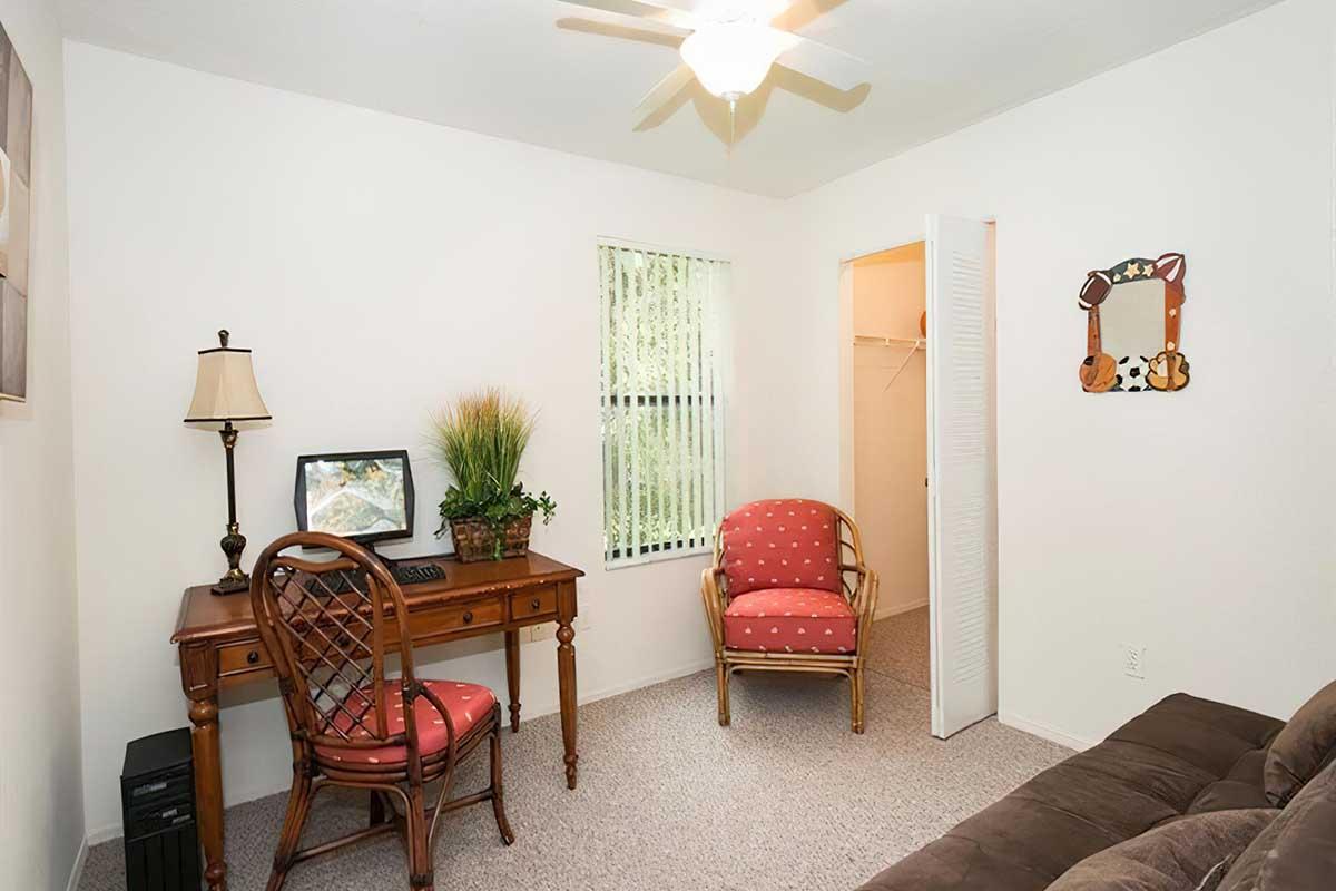 A cozy room featuring a desk with a computer monitor, a chair, and a small lamp. There are two red upholstered chairs with polka dots, a potted plant, and a wall mirror. A closet door is slightly ajar, and a window with blinds allows natural light to enter. The carpet is light-colored, creating a warm atmosphere.
