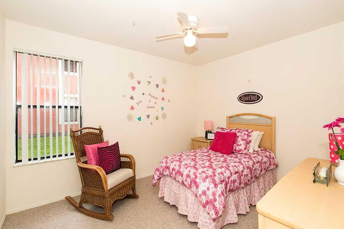 A cozy bedroom featuring a bed with floral bedding, pink accent pillows, and a decorative rocking chair. The walls are adorned with wall art, and there's a dresser and a window with blinds allowing natural light to enter the space.