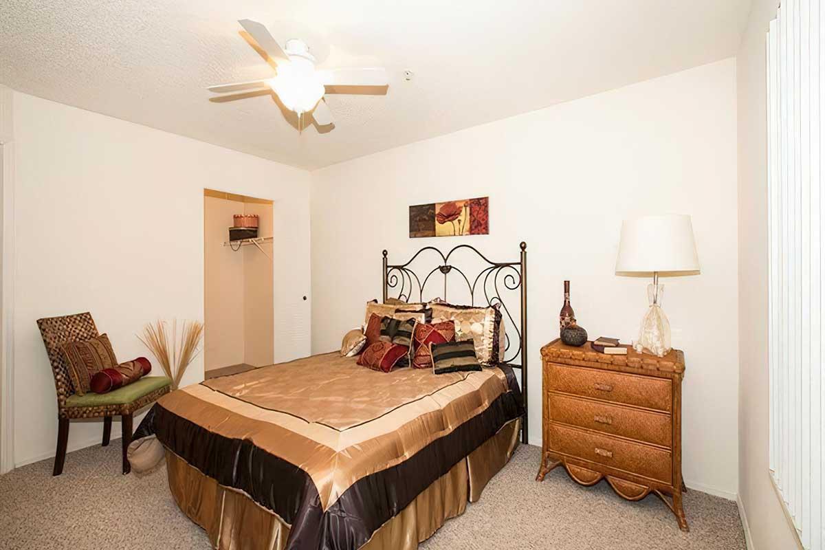 A cozy bedroom featuring a queen-sized bed with a decorative gold and brown comforter, accent pillows, and a wrought iron headboard. There's a bedside lamp on a wooden nightstand, a chair with decorative cushions, and a small closet space in the background. The walls are painted light, and a ceiling fan is visible.