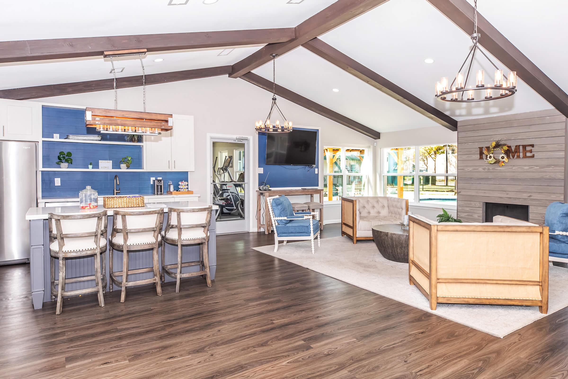 Modern living room and kitchen area featuring an open layout. The space includes a large kitchen island with bar stools, a dining area, comfortable seating options like armchairs, and large windows that provide natural light. The decor incorporates a mix of rustic and contemporary styles, with exposed beams and stylish lighting.