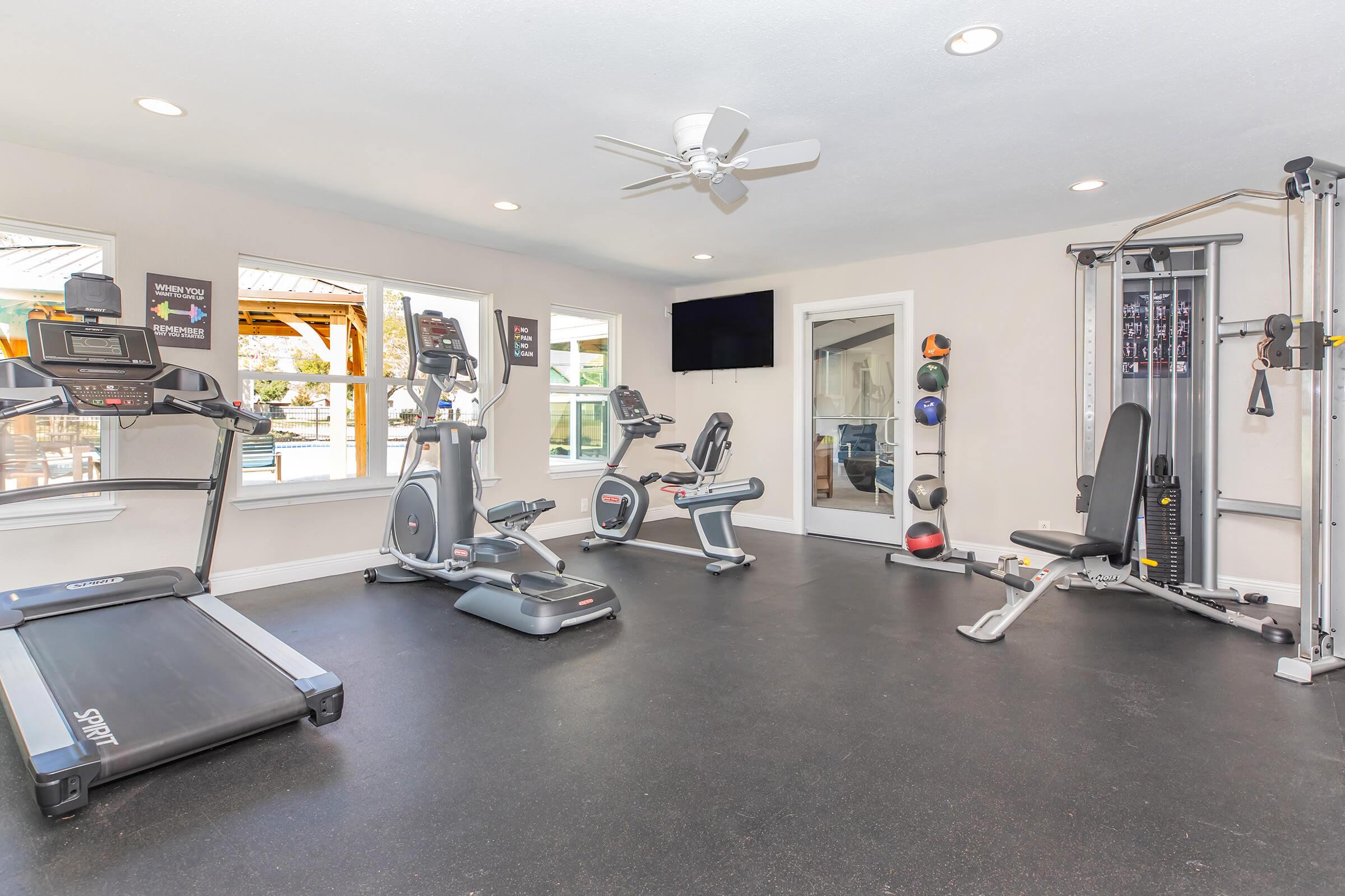 A well-lit fitness room featuring a treadmill, stationary bike, elliptical machine, and a multi-gym strength training machine. Large windows provide natural light, and there are mirrors on the wall. The space is designed for workout activities, with a ceiling fan and a television mounted for entertainment.