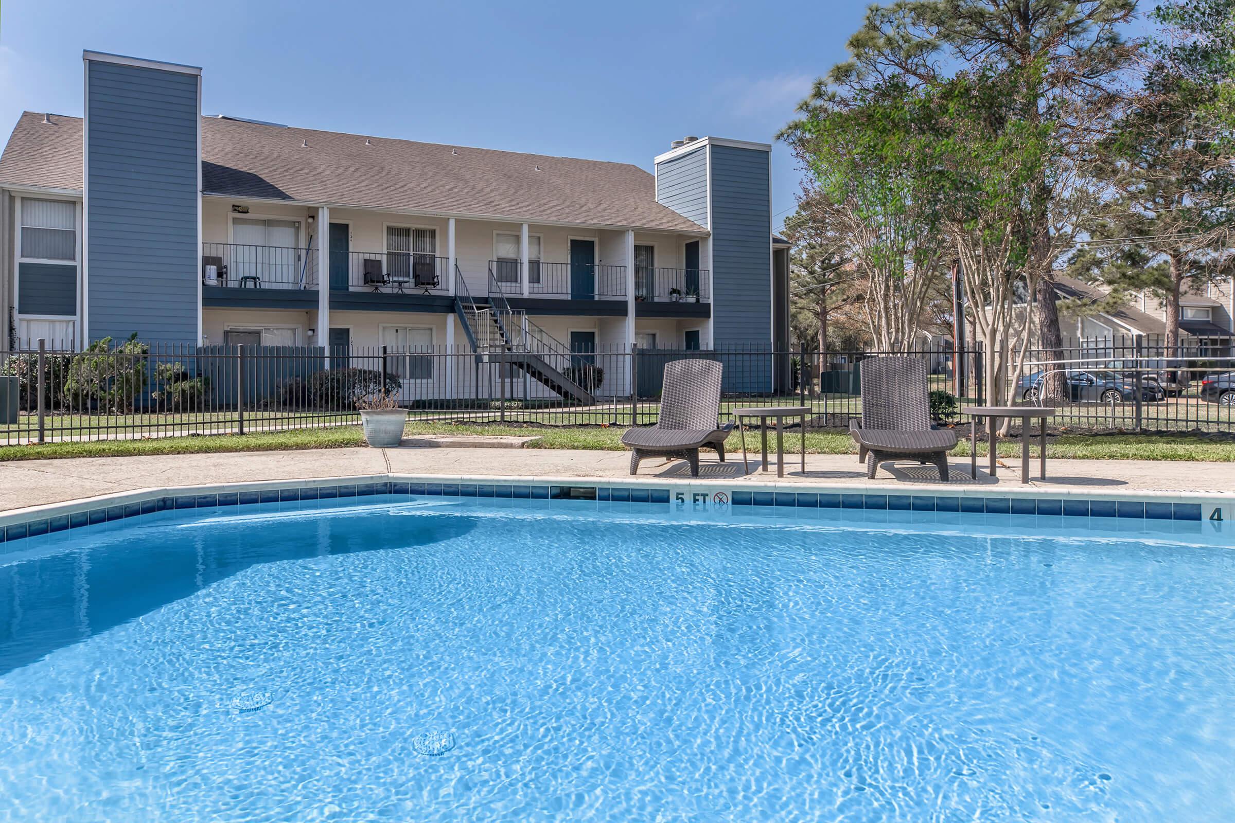 A clear outdoor swimming pool with a shallow area marked "5 FT" in front of a two-story apartment building. There are two lounge chairs near the pool, surrounded by a fenced area with trees in the background under a blue sky.