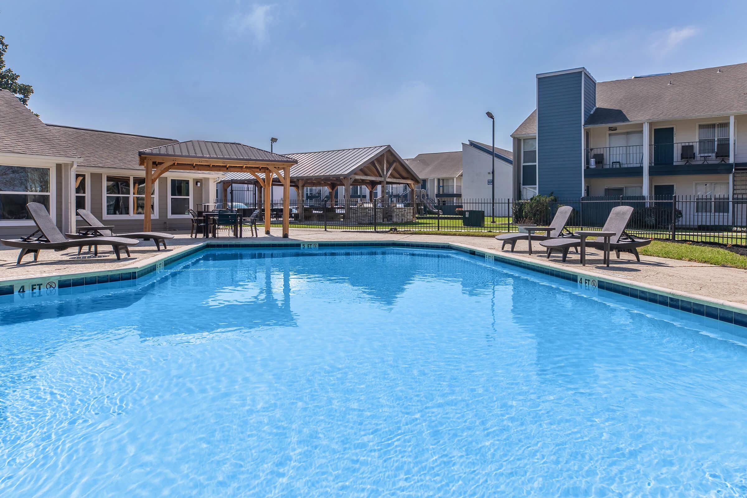 A refreshing blue swimming pool surrounded by lounge chairs, with shaded seating areas in the background. Nearby, buildings and a green lawn contribute to a relaxing outdoor atmosphere. The sky is clear, enhancing the inviting ambiance of the pool area.
