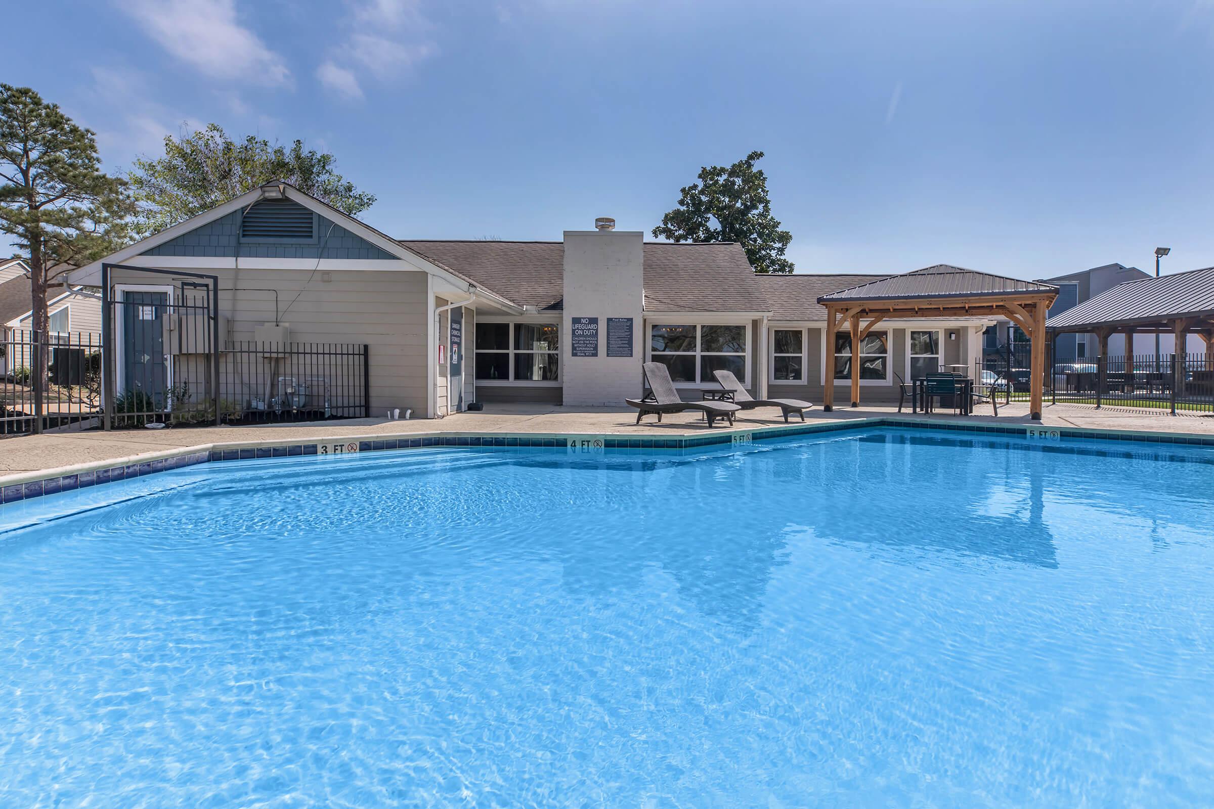 A clear blue swimming pool surrounded by a patio with lounge chairs. In the background, there is a building with large windows and a covered area. Trees are visible around the property under a bright blue sky.