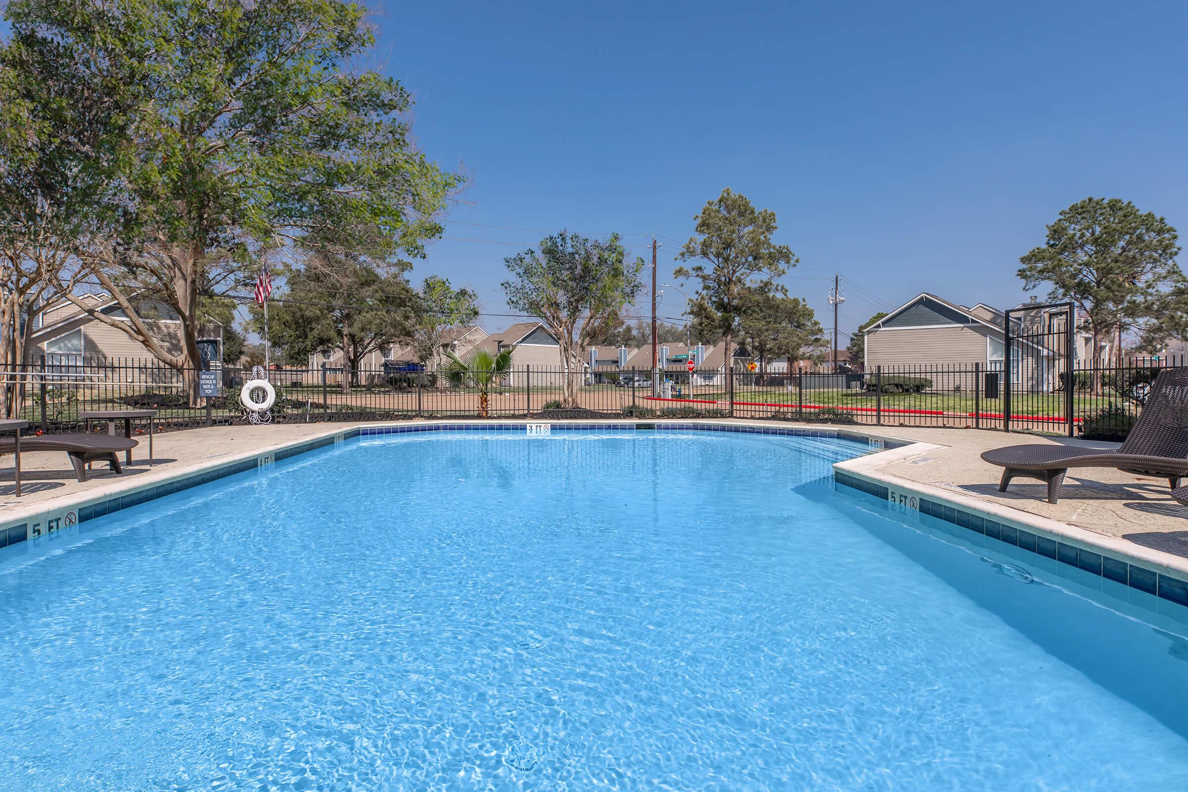 A clear blue swimming pool surrounded by lounge chairs, with a manicured landscape and trees in the background. Nearby, residential buildings can be seen, and the sky is bright and sunny. Safety equipment, like a life ring, is visible near the pool area.
