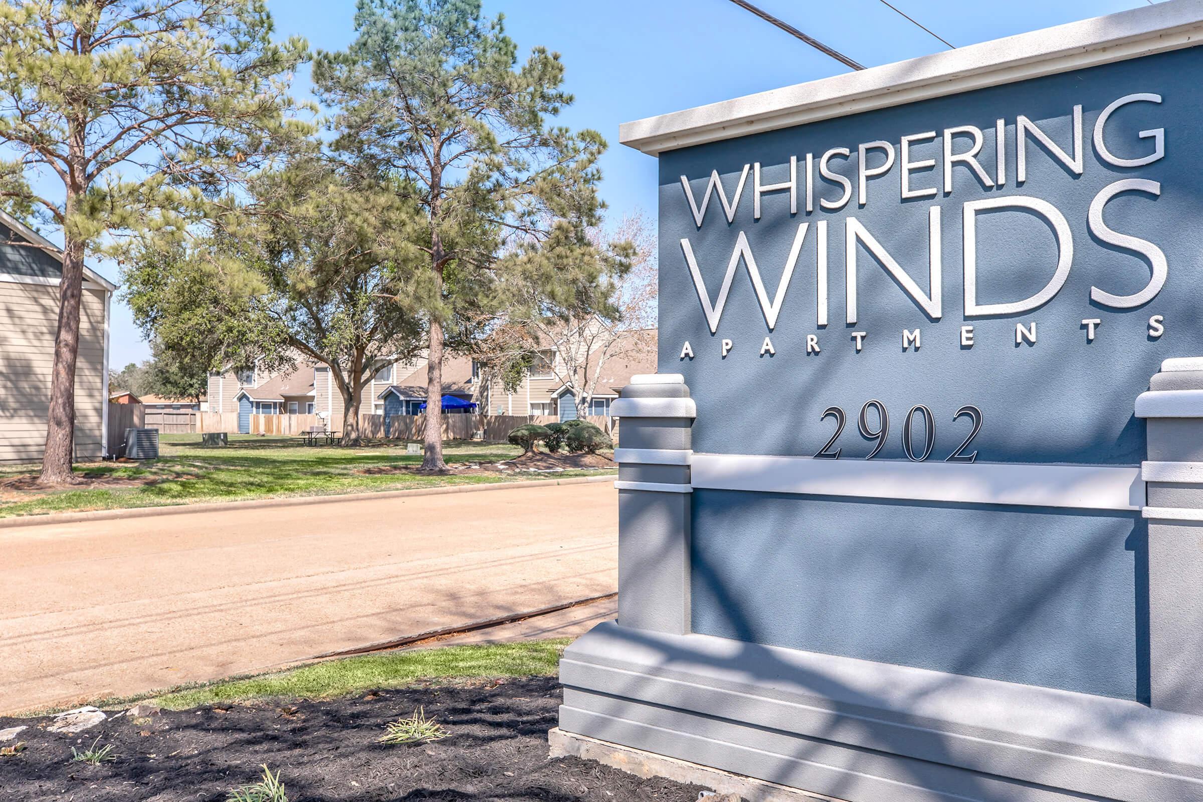 Sign for Whispering Winds Apartments featuring the name prominently displayed. The sign is set against a backdrop of trees and a clear blue sky, with a road visible in the foreground. The apartment complex is located at 2902.