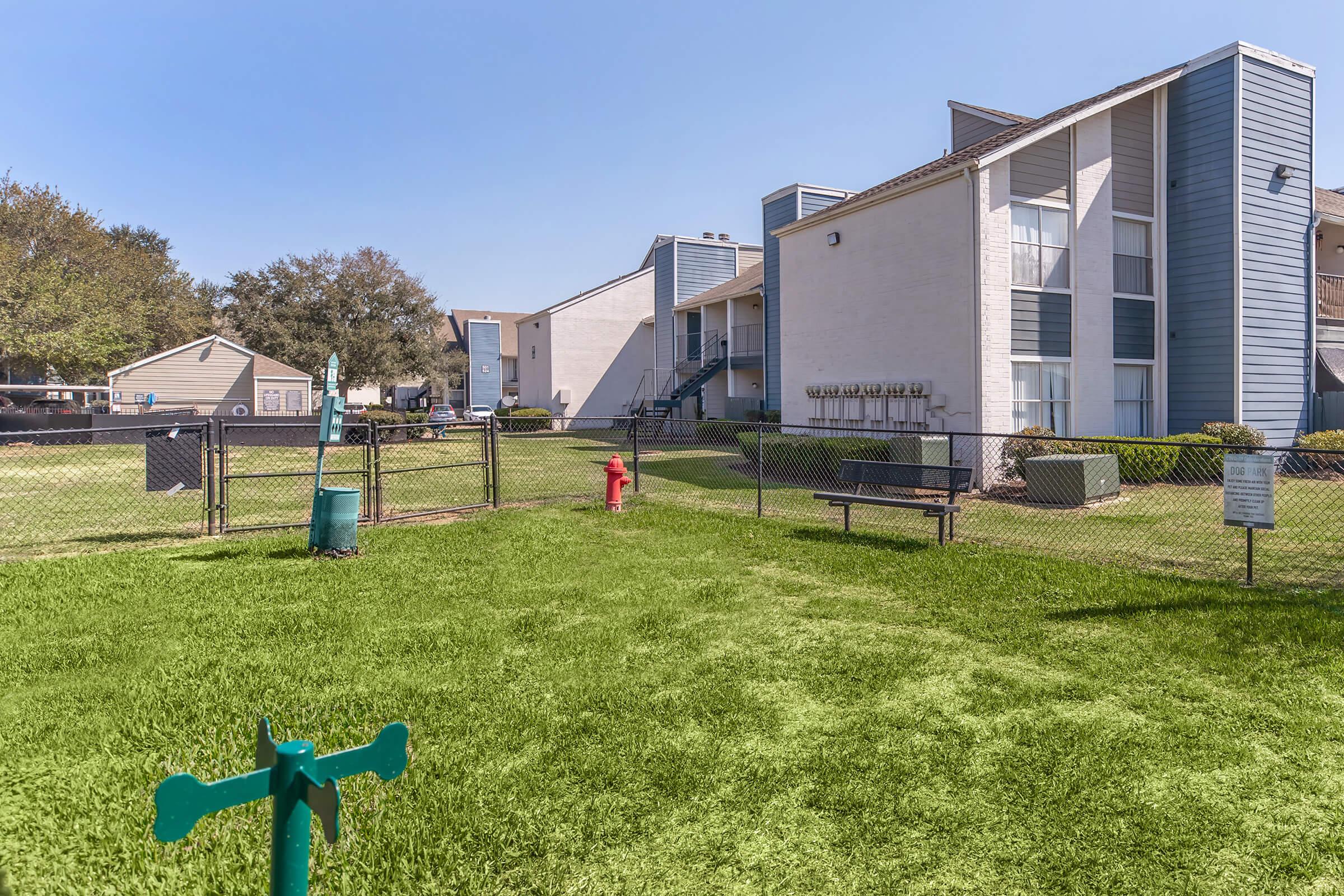 A well-maintained grassy area with a green hydrant and a blue water source is fenced off, surrounded by two multi-story residential buildings. The scene shows a clear sky and landscaped surroundings, suggesting a pleasant environment for residents and pets.