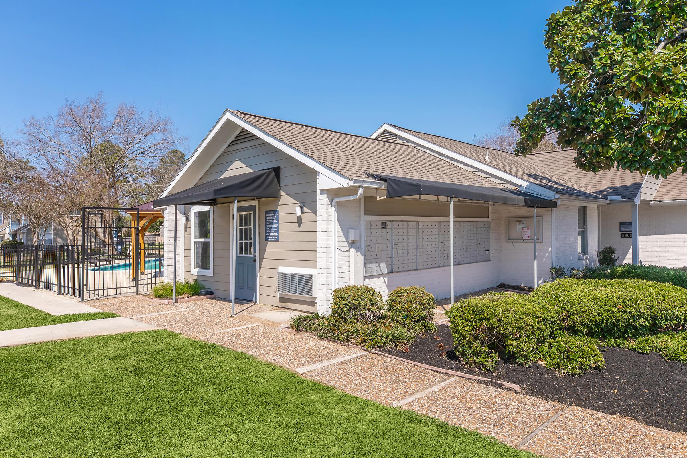 A well-maintained residential building with a light gray façade and a dark gray door. It features a covered entryway and landscaped gardens with neatly trimmed bushes. A swimming pool is visible in the background, and the sky is clear and blue.