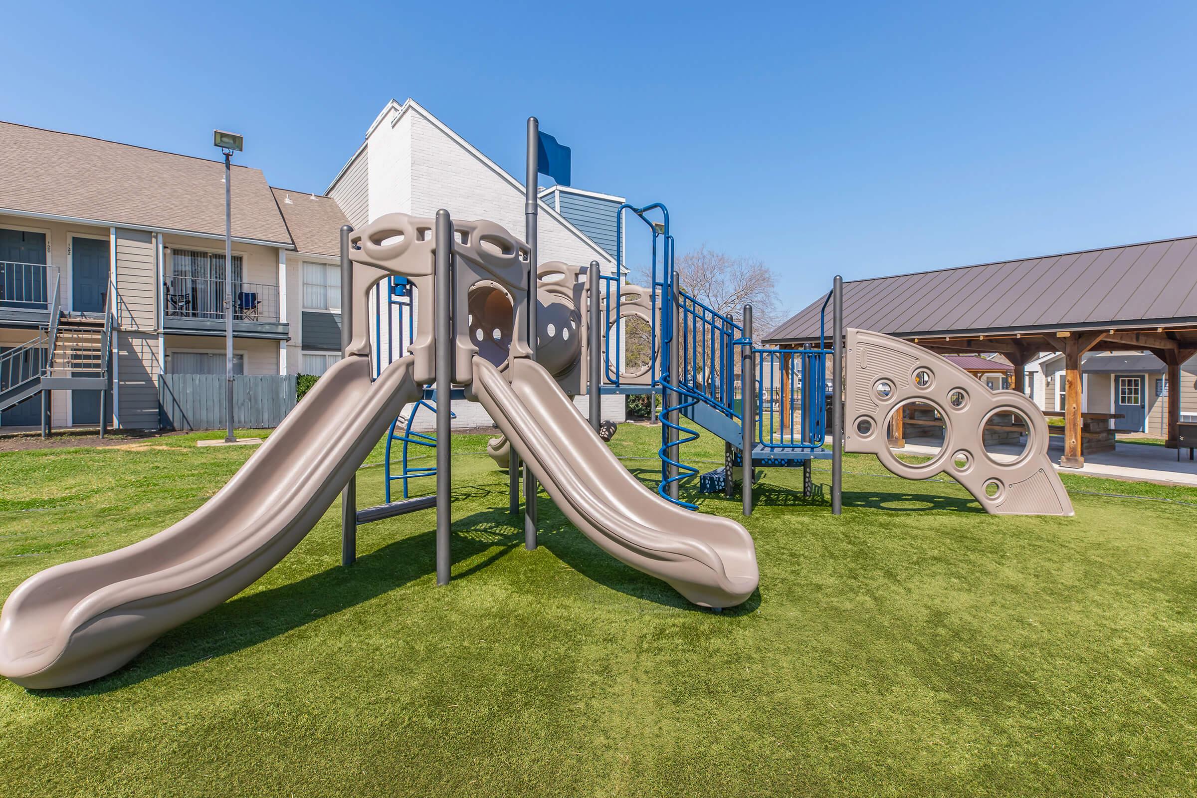 A playground featuring two tan slides, climbing structures, and a play area on artificial grass. Background includes a building with multiple units and a covered picnic area. The sky is clear and blue, creating a bright and inviting atmosphere.