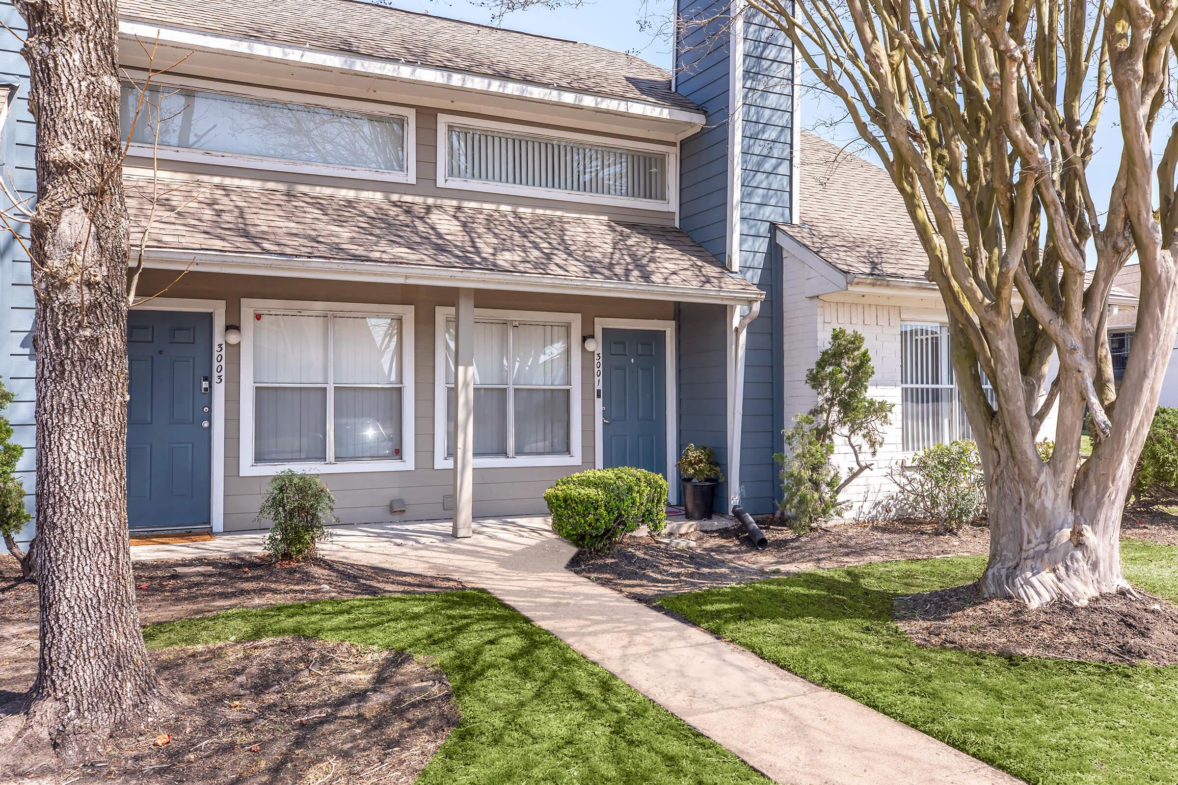 A suburban townhouse with a blue and gray exterior, featuring two front doors, large windows, and manicured landscaping. A clear path leads to the entrance, bordered by green lawn and shrubs, with two trees framing the front. The scene is bright and inviting, suggesting a well-maintained community.