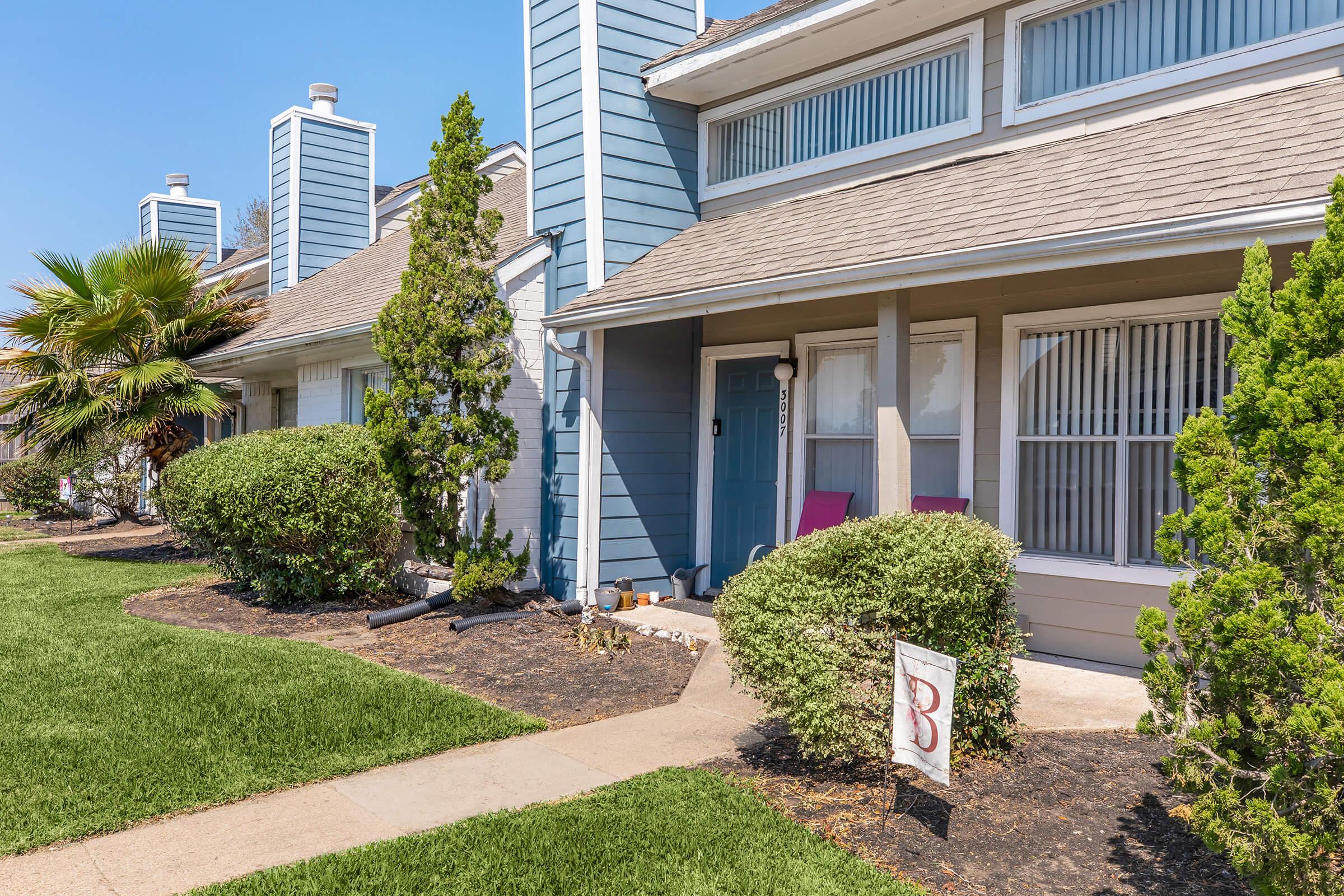 A well-maintained two-story blue townhouse with a landscaped front yard. The entrance features a door adorned with a number, flanked by tall shrubs and palm trees. Two purple chairs are placed on the porch, and a sign is positioned on the lawn. The clear blue sky completes the inviting atmosphere.
