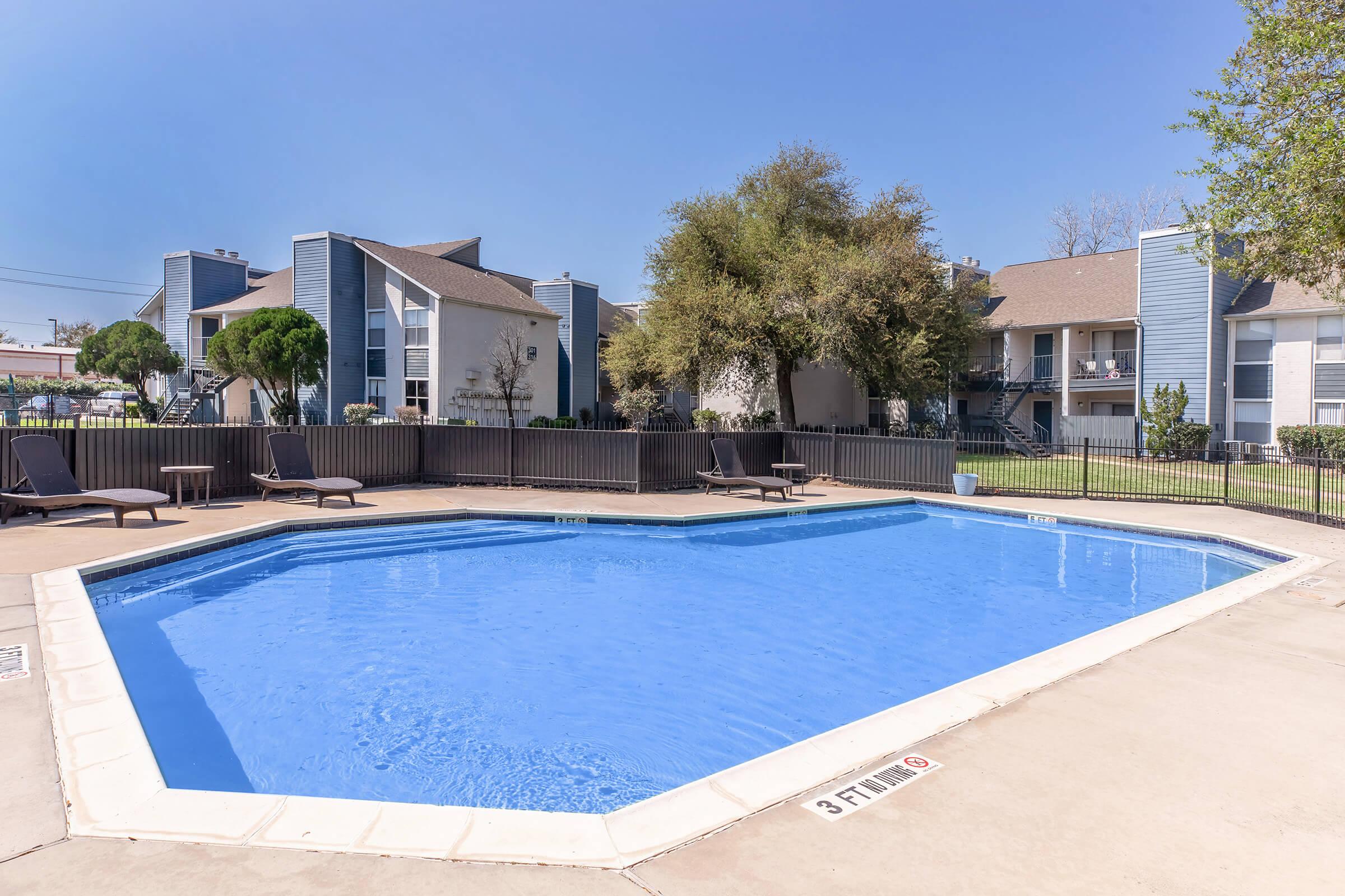 A clear blue swimming pool surrounded by a concrete deck and lounge chairs. In the background, there are several multi-story residential buildings with balconies and green trees. The sky is bright and sunny, indicating a pleasant day.