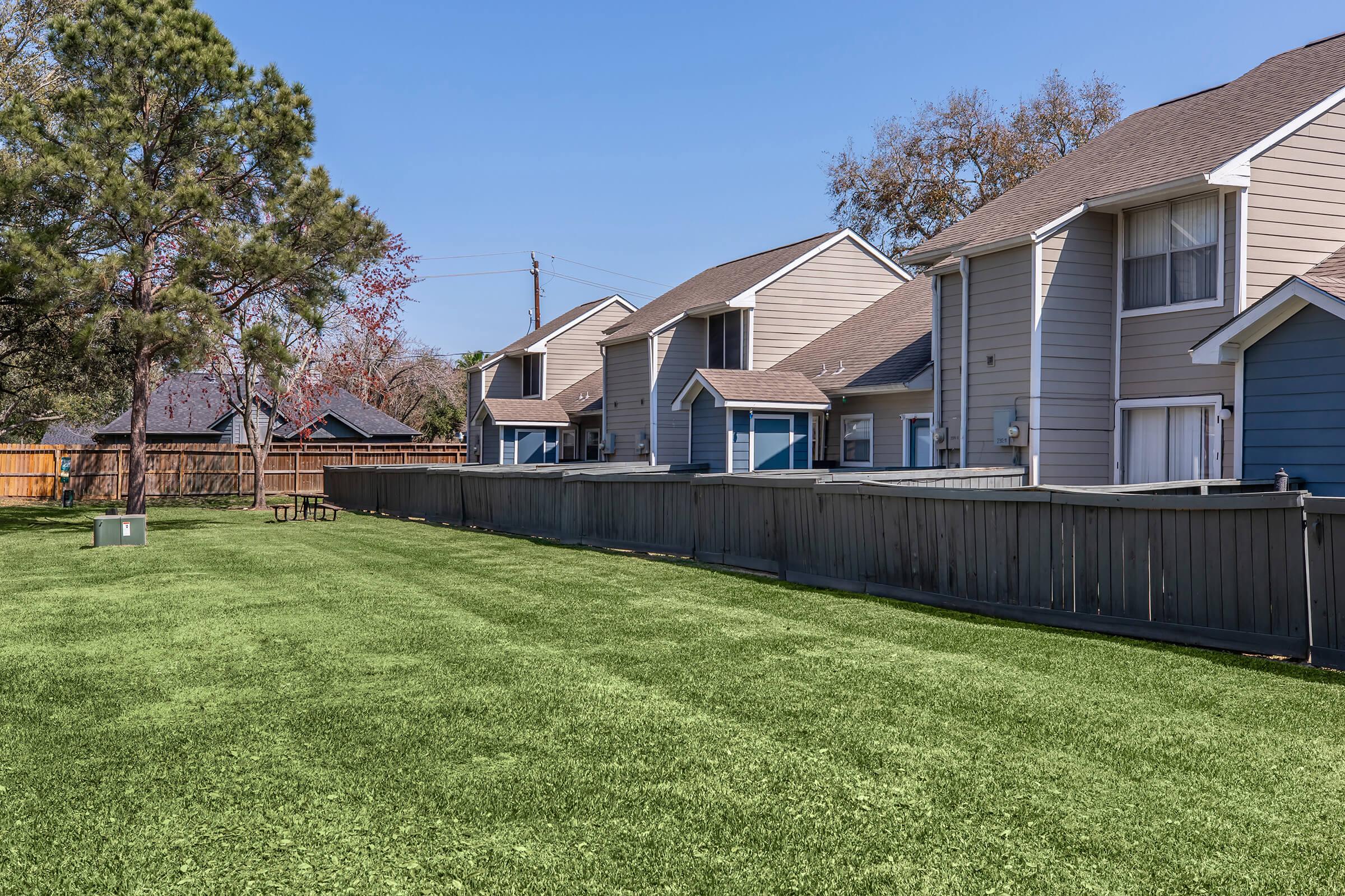 A well-maintained grassy backyard with two residential buildings on the right. A wooden fence delineates the property, and a tree provides some shade. The sky is clear and blue, indicating a sunny day.