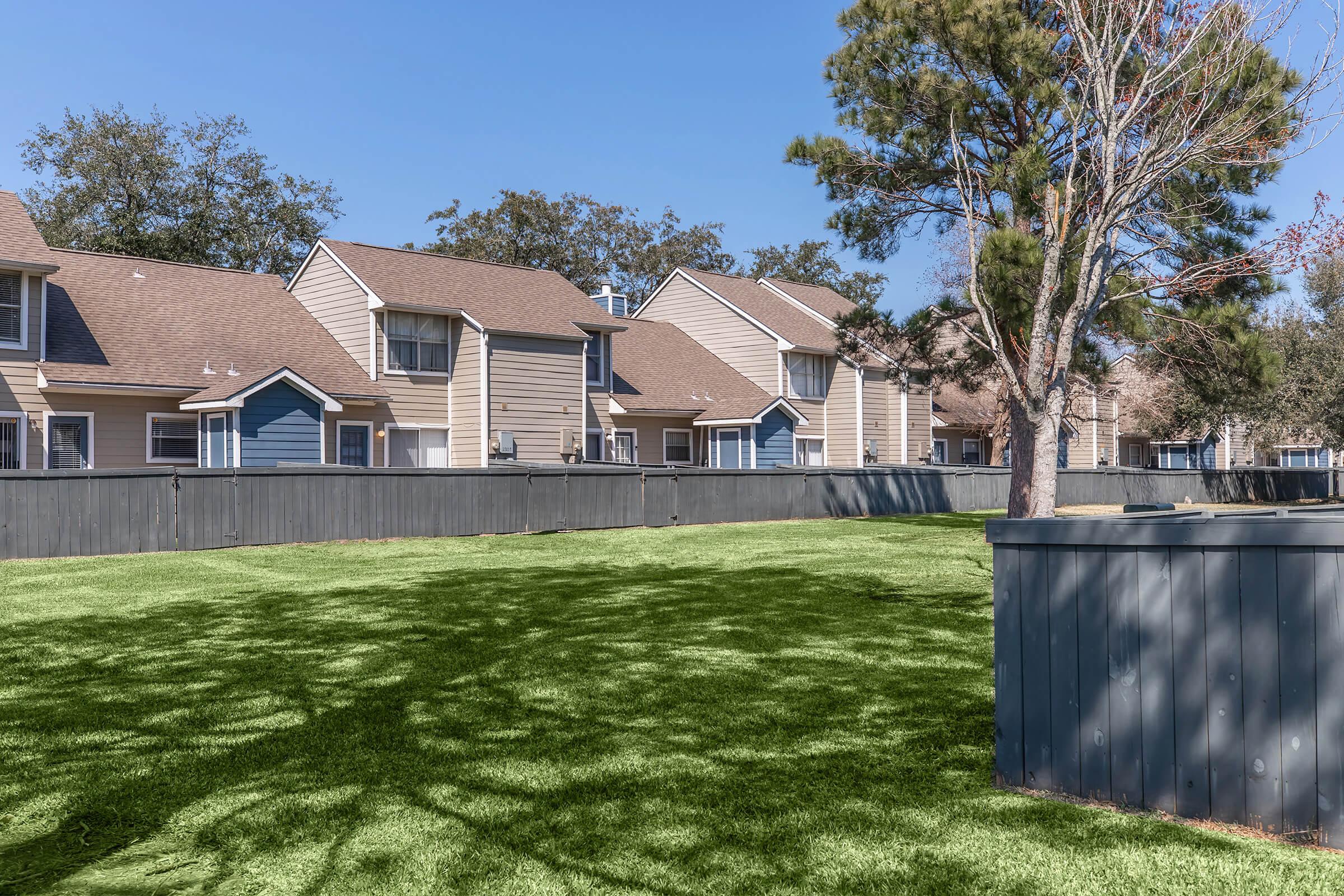 A row of two-story residential buildings behind a wooden fence, with a well-maintained green lawn in the foreground. Tall trees provide shade, and the bright blue sky adds to the serene outdoor ambiance. The setting appears calm and inviting, suitable for family living.