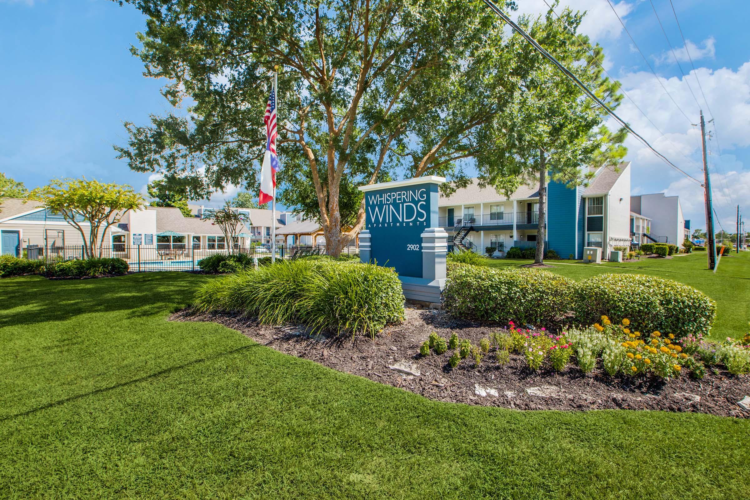 A well-maintained outdoor area featuring a large green lawn, colorful flower beds, and a sign that reads "Whispering Winds." In the background, there are buildings with a modern design, along with trees and flagpoles displaying flags. The sky is partly cloudy, suggesting a pleasant day.