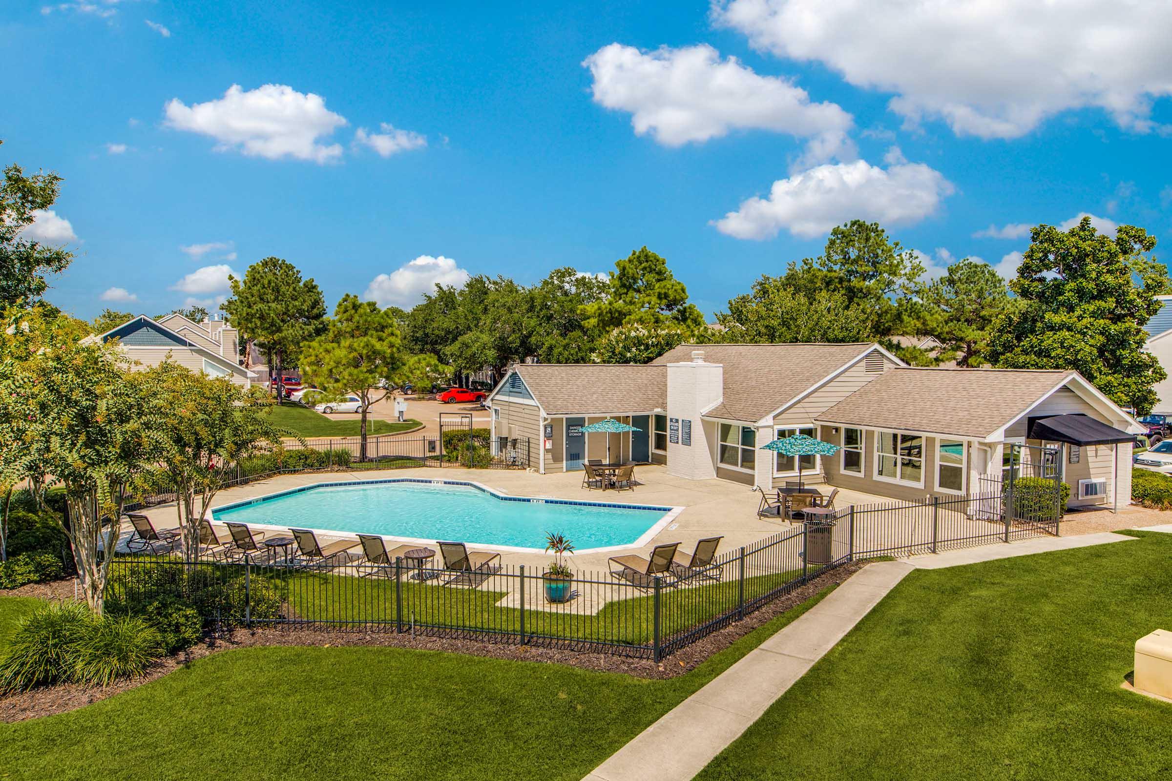 Aerial view of a residential pool area featuring a clear blue swimming pool surrounded by lounge chairs and umbrellas. A clubhouse is nearby, set in a landscaped environment with trees and well-maintained grass. The sky is bright with fluffy clouds, creating a serene and inviting atmosphere.