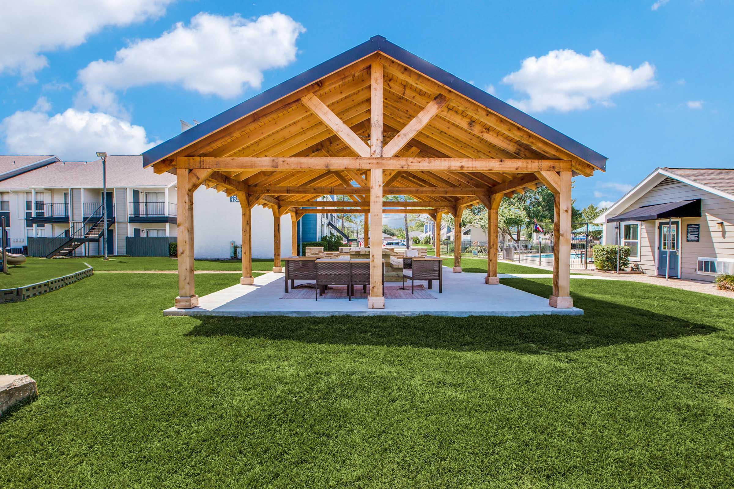 A spacious outdoor pavilion with a wooden frame and a gabled roof, surrounded by lush green grass. Under the pavilion, there are several patio chairs and a table arranged for seating. In the background, there are residential buildings and a bright blue sky with scattered clouds.