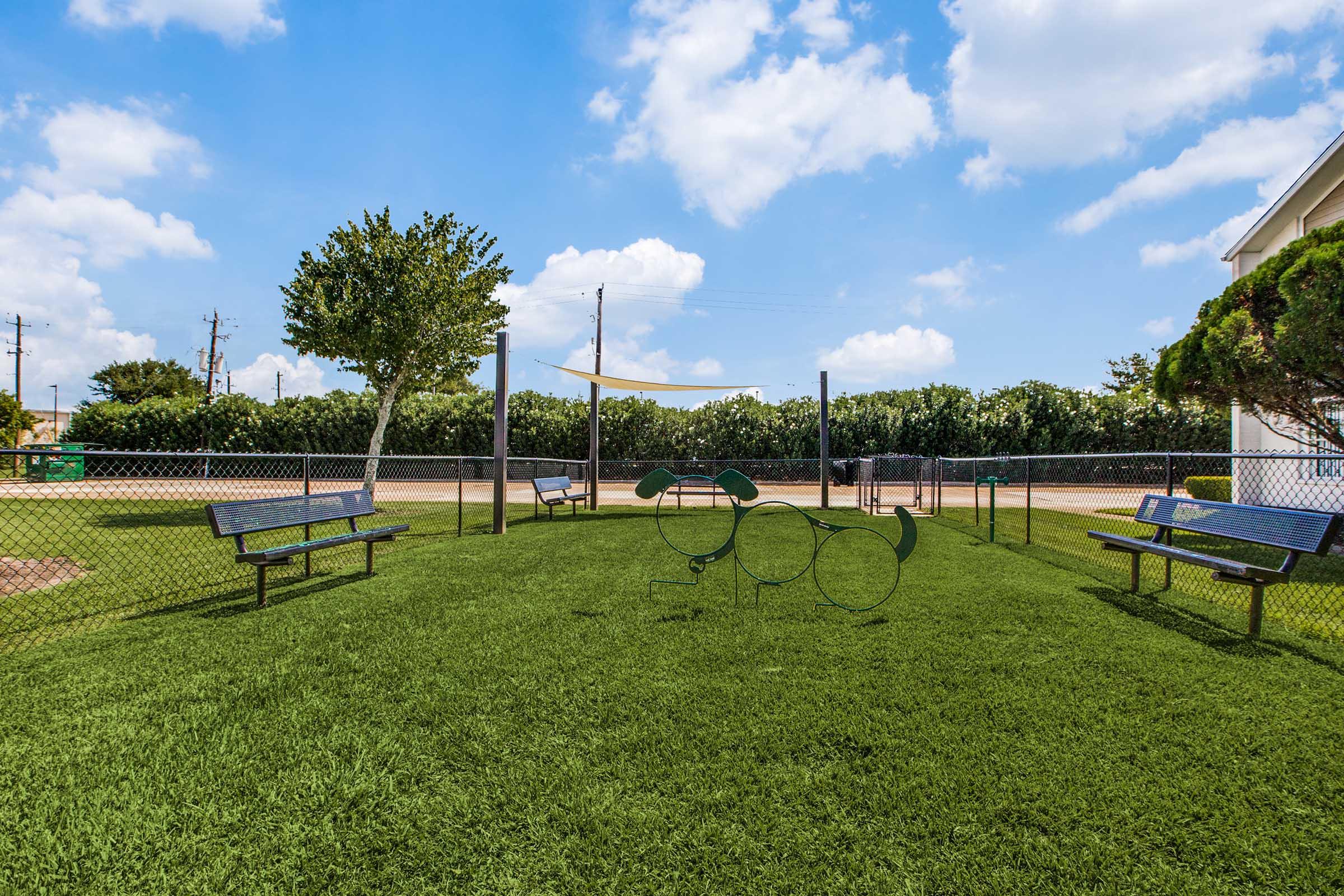 A grassy playground area featuring green benches and unique metal sculptures resembling animals. In the background, there's a fence and a shaded area, surrounded by trees and a clear blue sky with clouds.