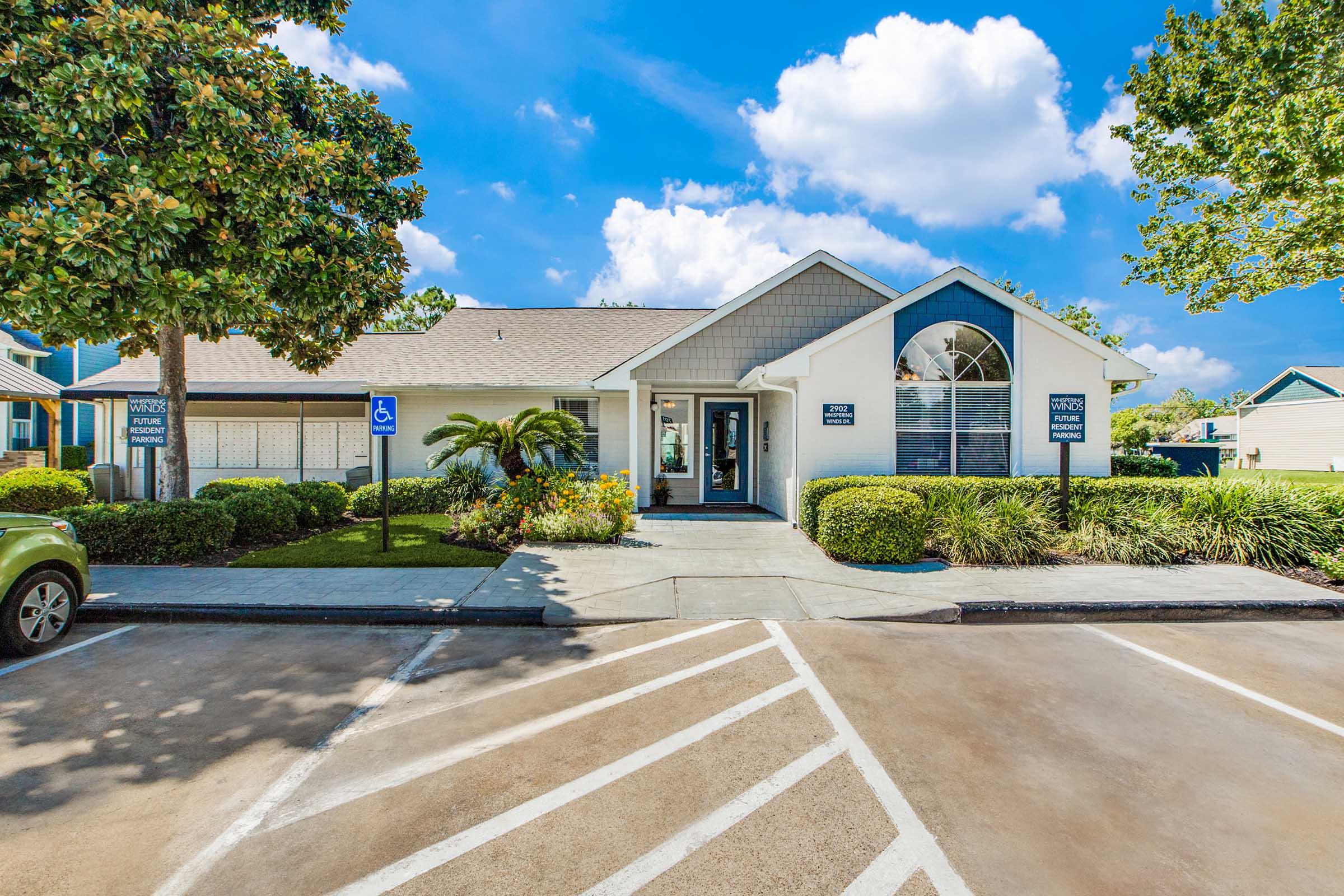 A modern office building surrounded by greenery and landscaping. The structure features a pitched roof, large windows, and accessible parking spots. Bright blue sky with a few clouds enhances the inviting atmosphere of the location.