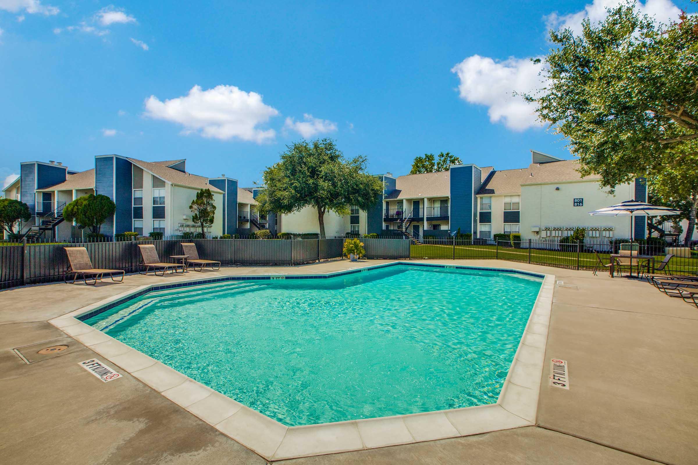 A clear, inviting swimming pool surrounded by lounge chairs, situated in a residential area with blue and white apartment buildings in the background under a bright blue sky with a few clouds. Green trees and manicured grass enhance the pleasant outdoor setting.