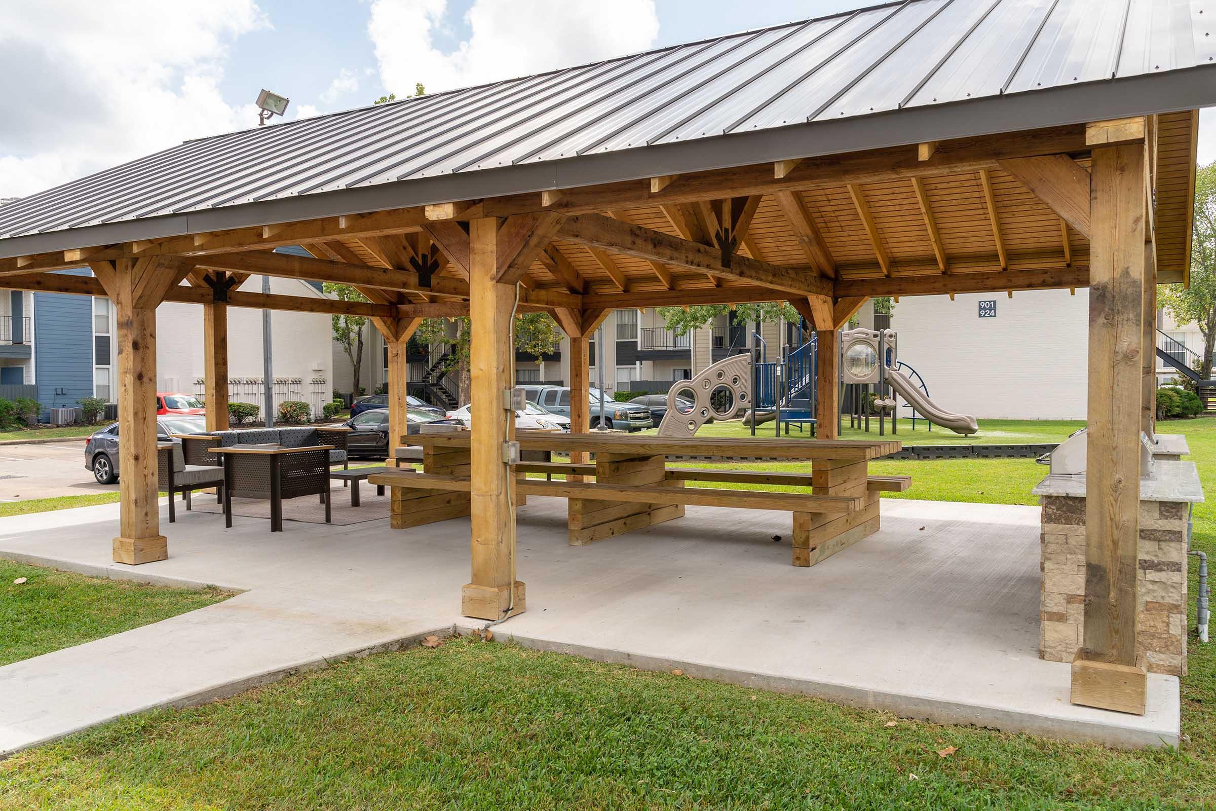 Covered picnic area with wooden beams, benches, and tables, situated on a grassy lawn. In the background, a playground featuring slides and climbing structures is visible. The sky is partly cloudy, enhancing the outdoor setting.