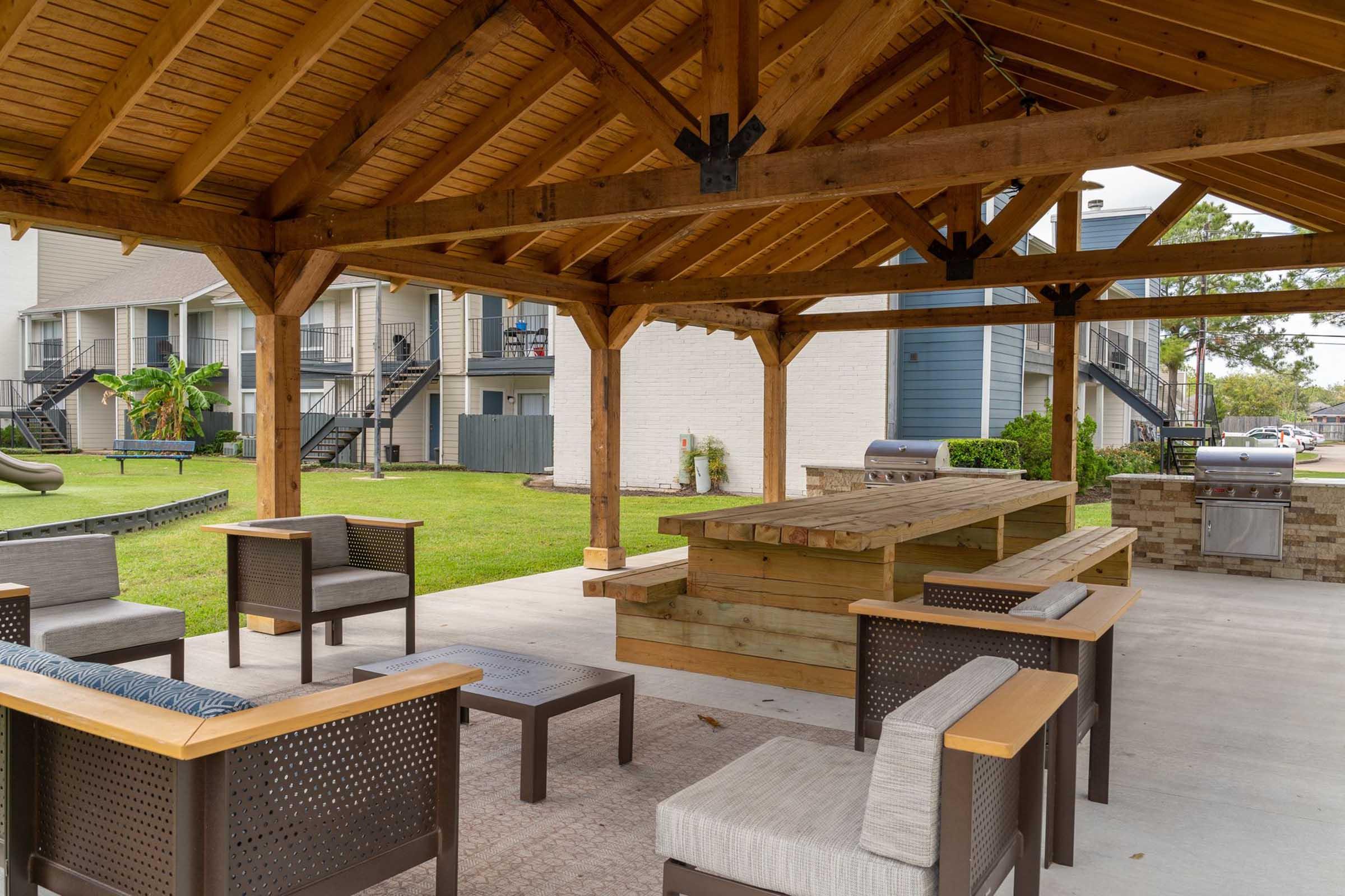A spacious outdoor pavilion with a wooden roof and seating area. There are several chairs and a large wooden table, along with a barbecue grill. Surrounding greenery and buildings are visible in the background, creating a welcoming community space for gatherings and relaxation.