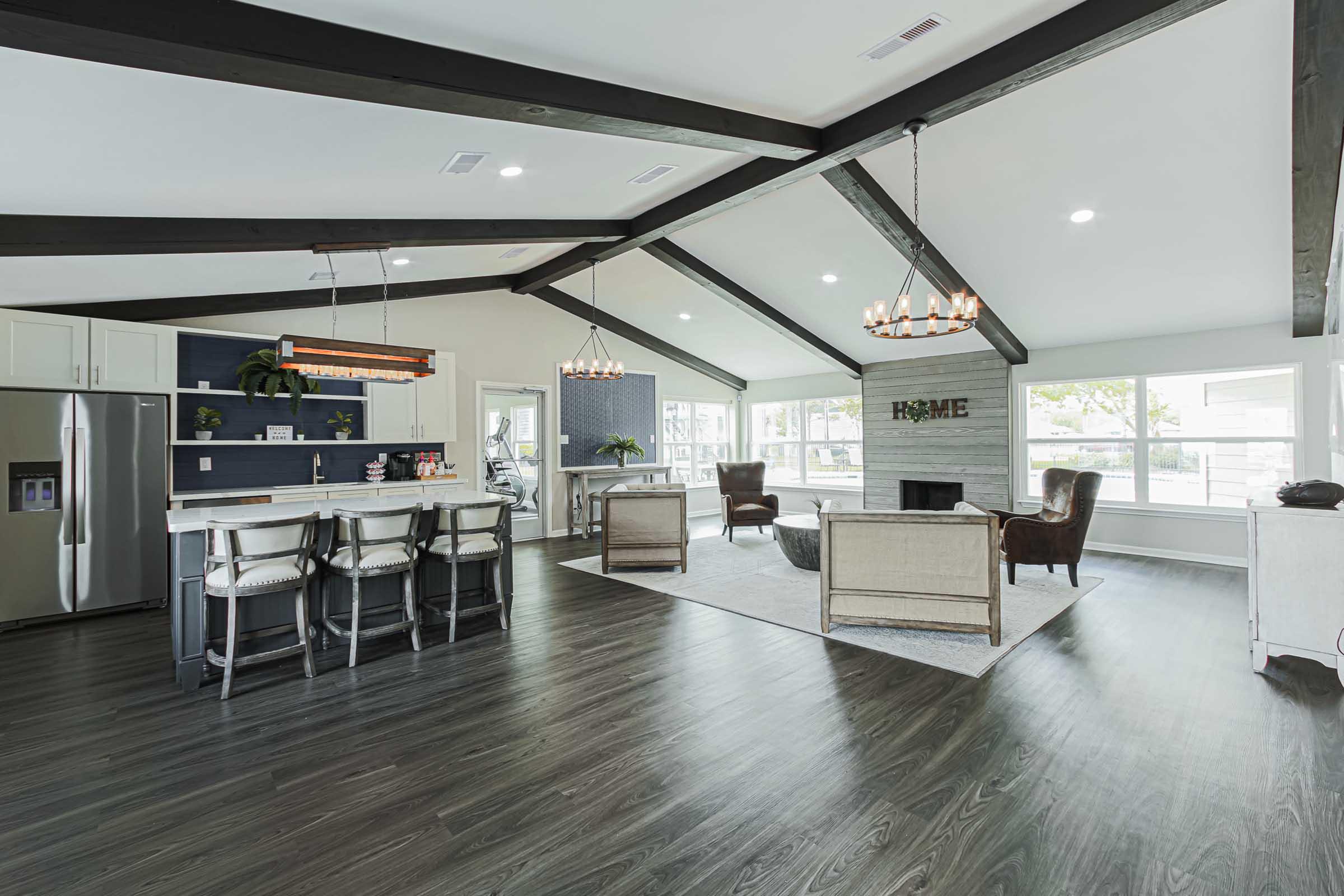 Spacious modern living area featuring an open floor plan. The kitchen includes a bar with seating, sleek appliances, and decorative shelving. A cozy sitting area with two armchairs, a fireplace, and large windows offers ample natural light. Neutral color palette and contemporary decor enhance the inviting atmosphere.