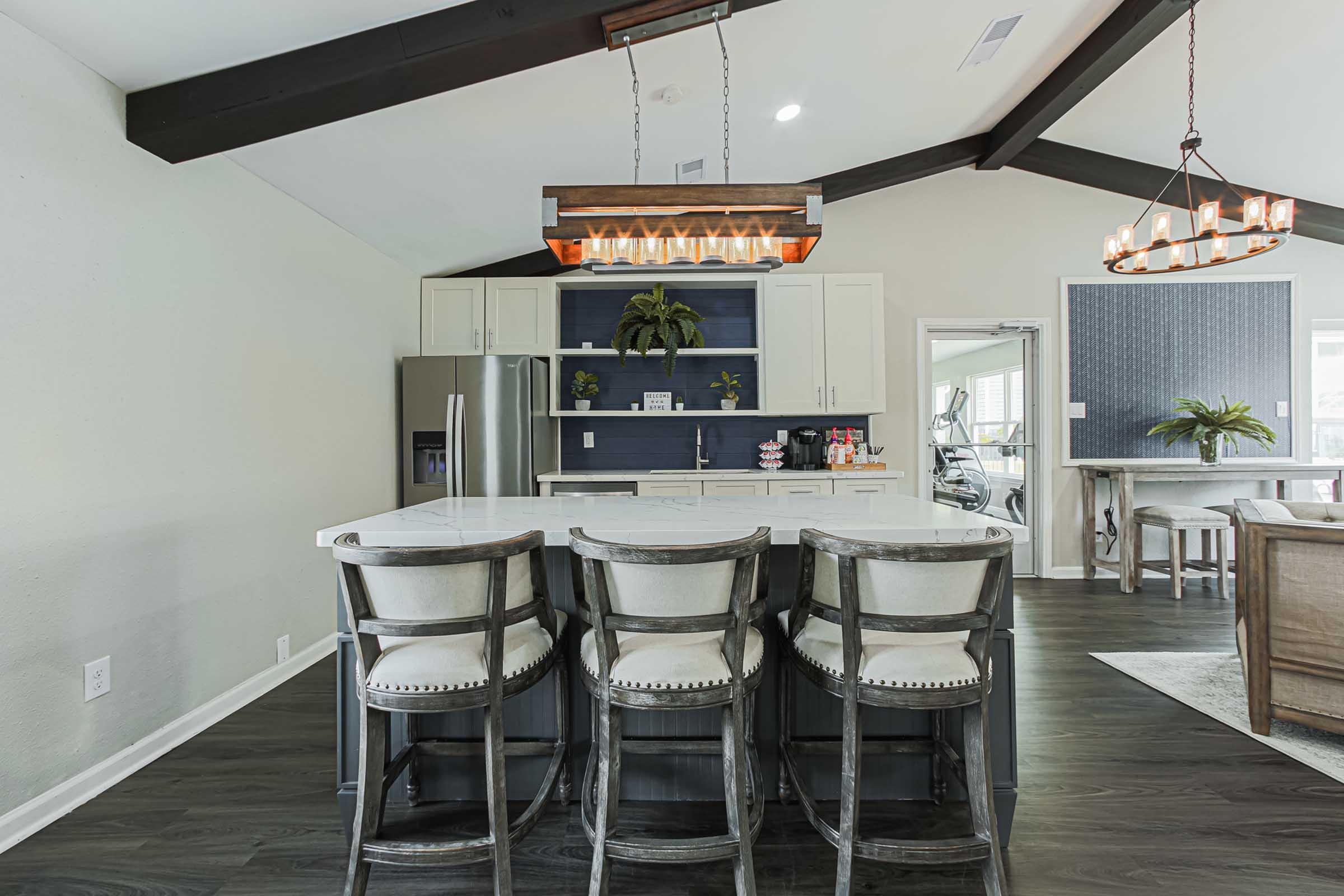 A modern kitchen with an island dining area featuring four high-backed bar stools. The space has dark wood beams on the ceiling, white cabinetry, and a stylish pendant light above the island. A dining area with a large table and decorative plant is visible in the background.