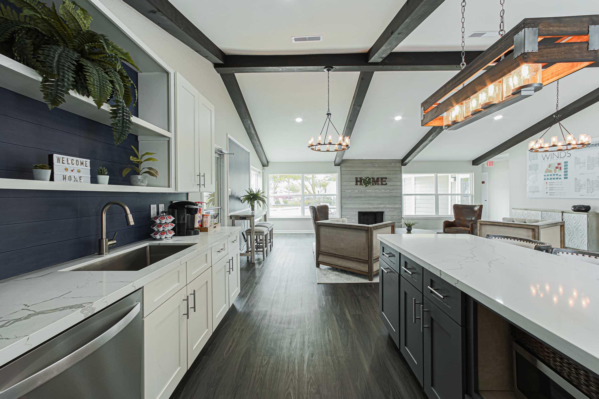 A modern kitchen featuring white and dark cabinetry, a spacious island with a marble countertop, and a stainless-steel sink. The room has exposed wooden beams, decorative plants, and a cozy living area visible in the background with a fireplace and large windows letting in natural light.