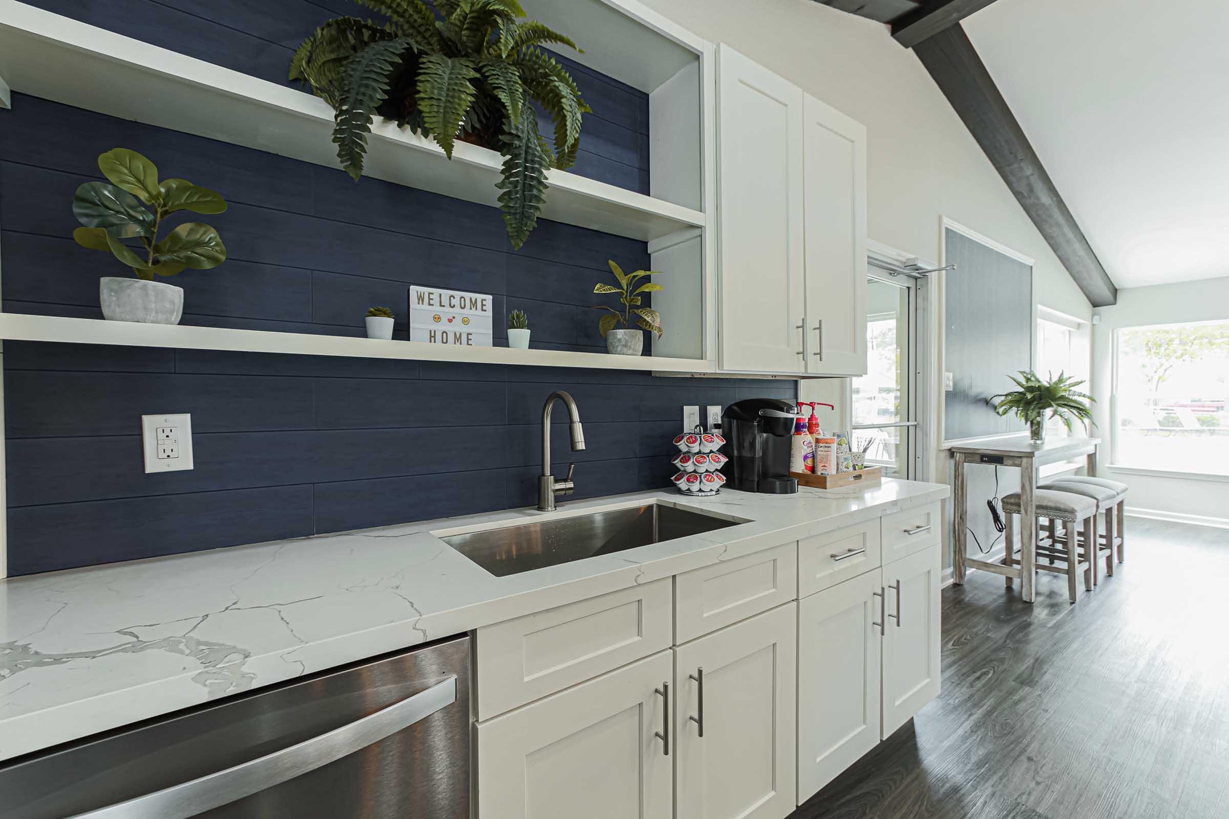A modern kitchen featuring white cabinetry, a marble countertop, and a stainless steel sink. Shelves display potted plants and a sign that reads "WELCOME HOME." To the right, there's a coffee maker and other kitchen accessories, with a bright and airy dining area in the background.
