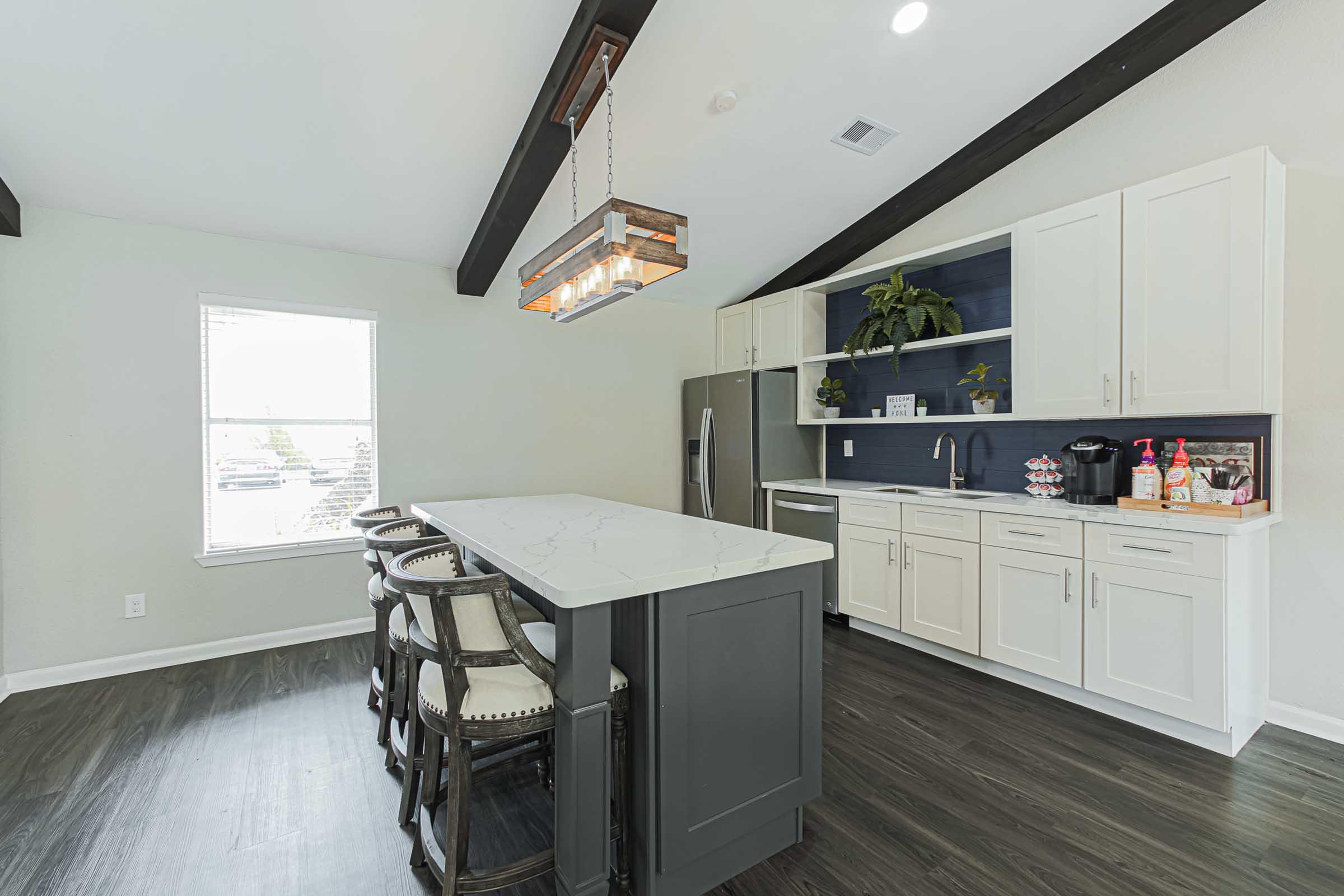 A modern kitchen featuring a large island with seating, white cabinetry, and dark wood beams. The space includes stainless steel appliances, a sink, and decorative plants. Natural light streams in through a window, creating a bright, open atmosphere.
