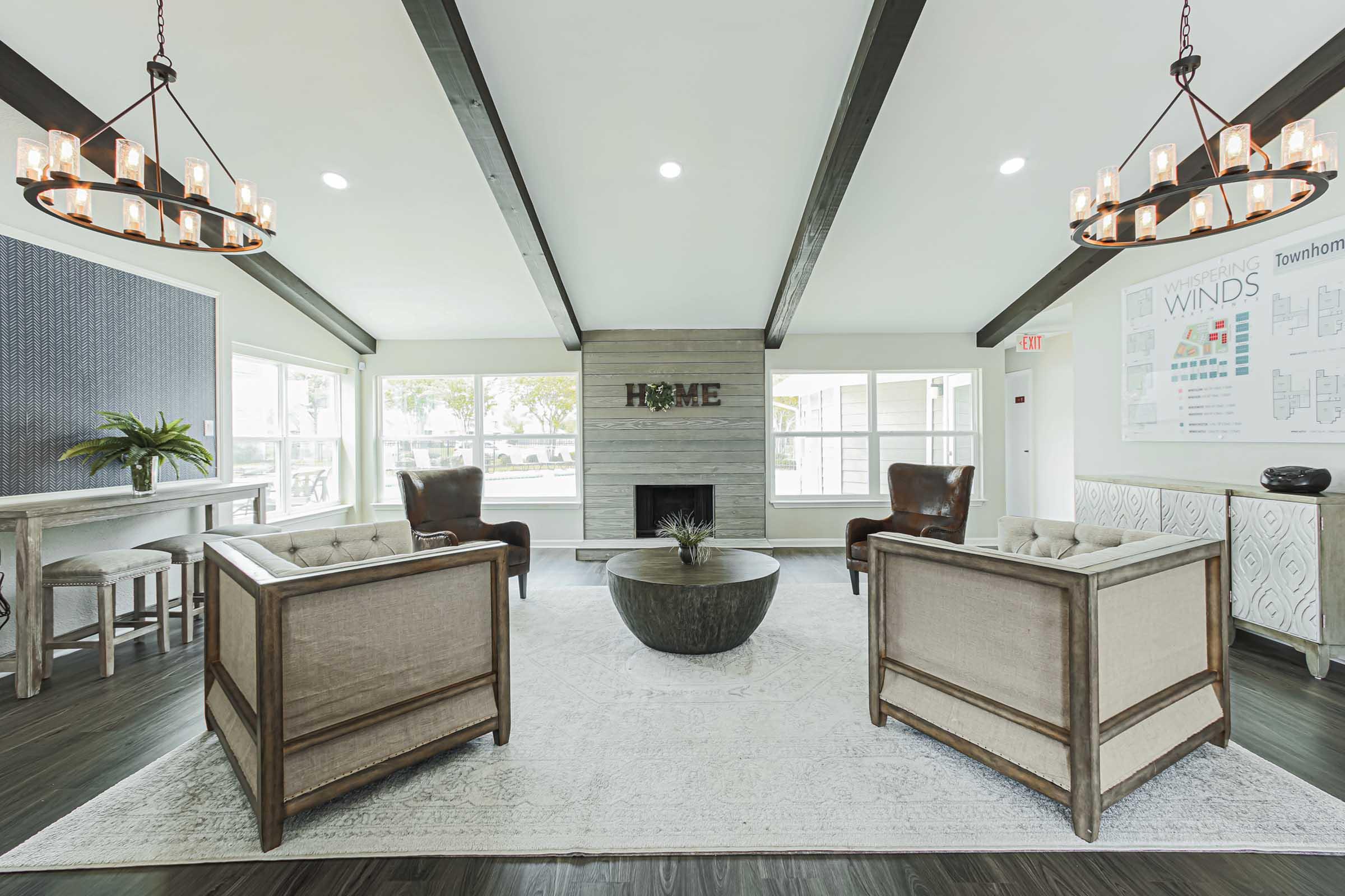 A modern living room featuring two elegant armchairs, a round coffee table, and a cozy fireplace. The space has large windows allowing natural light, with a neutral color palette and stylish light fixtures. A decorative plant adds a touch of greenery, while a wall displays information about townhomes.