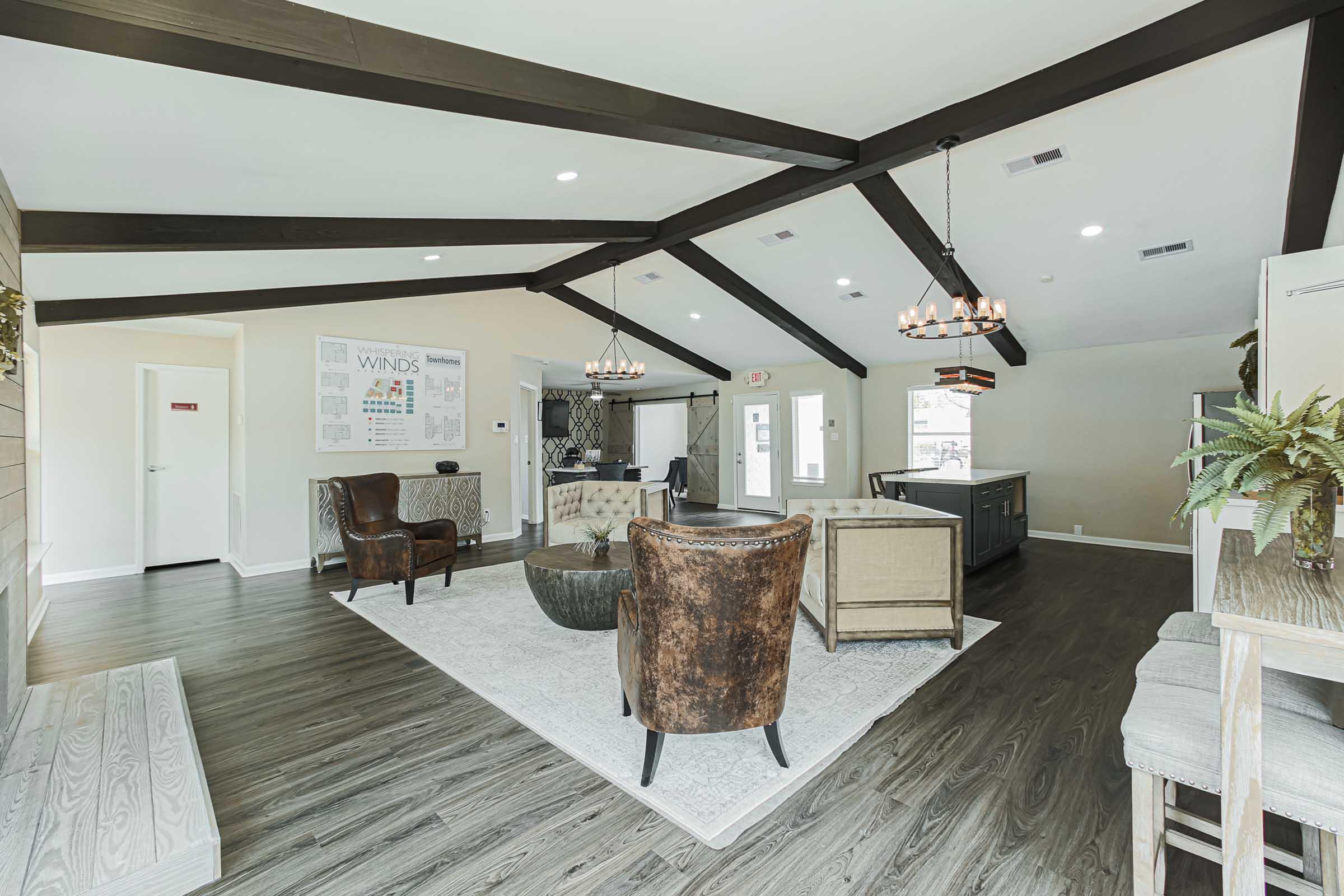 Spacious interior of a modern common area featuring exposed wooden beams, comfortable seating with leather armchairs, a round coffee table, and a welcoming reception desk. Brightly lit with large windows, the decor includes subtle greenery and a neutral color palette, creating a cozy yet stylish atmosphere.