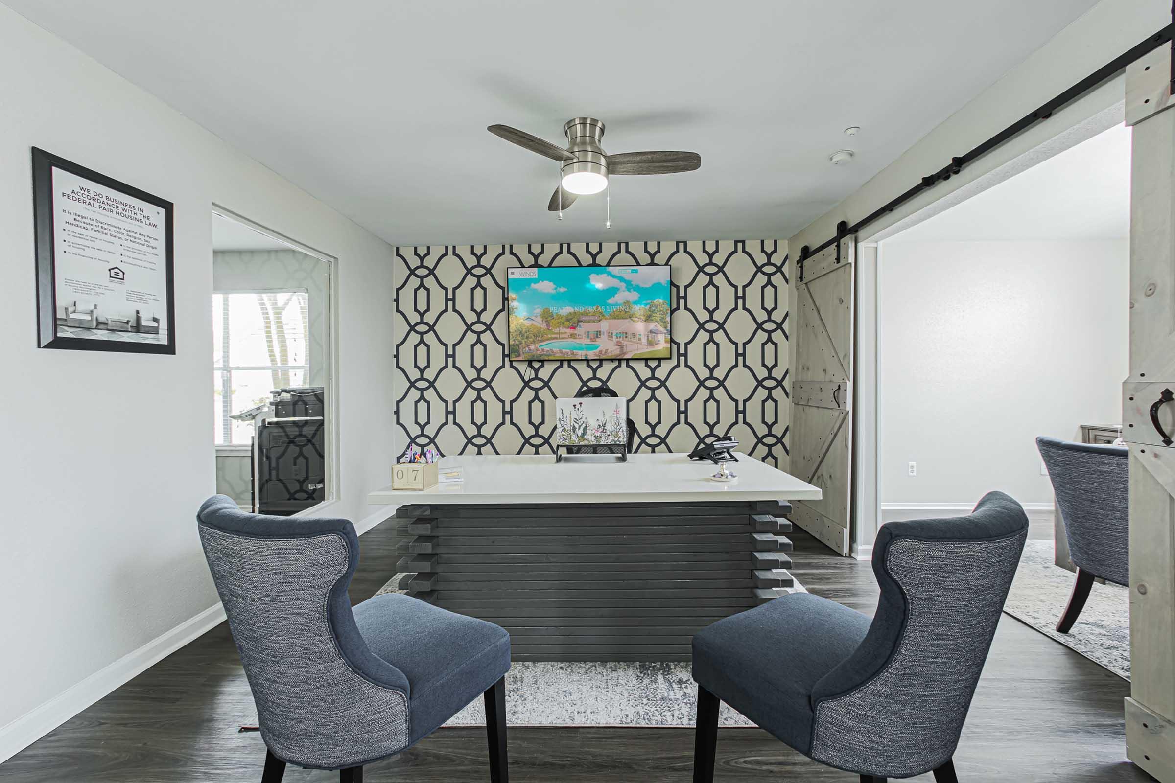 A modern office space featuring a sleek white desk with a decorative piece, two stylish gray chairs, and a decorative wall with a geometric pattern. A ceiling fan is visible above, and a colorful artwork is displayed on the wall, adding a touch of creativity to the environment. Natural light flows in from a nearby window.