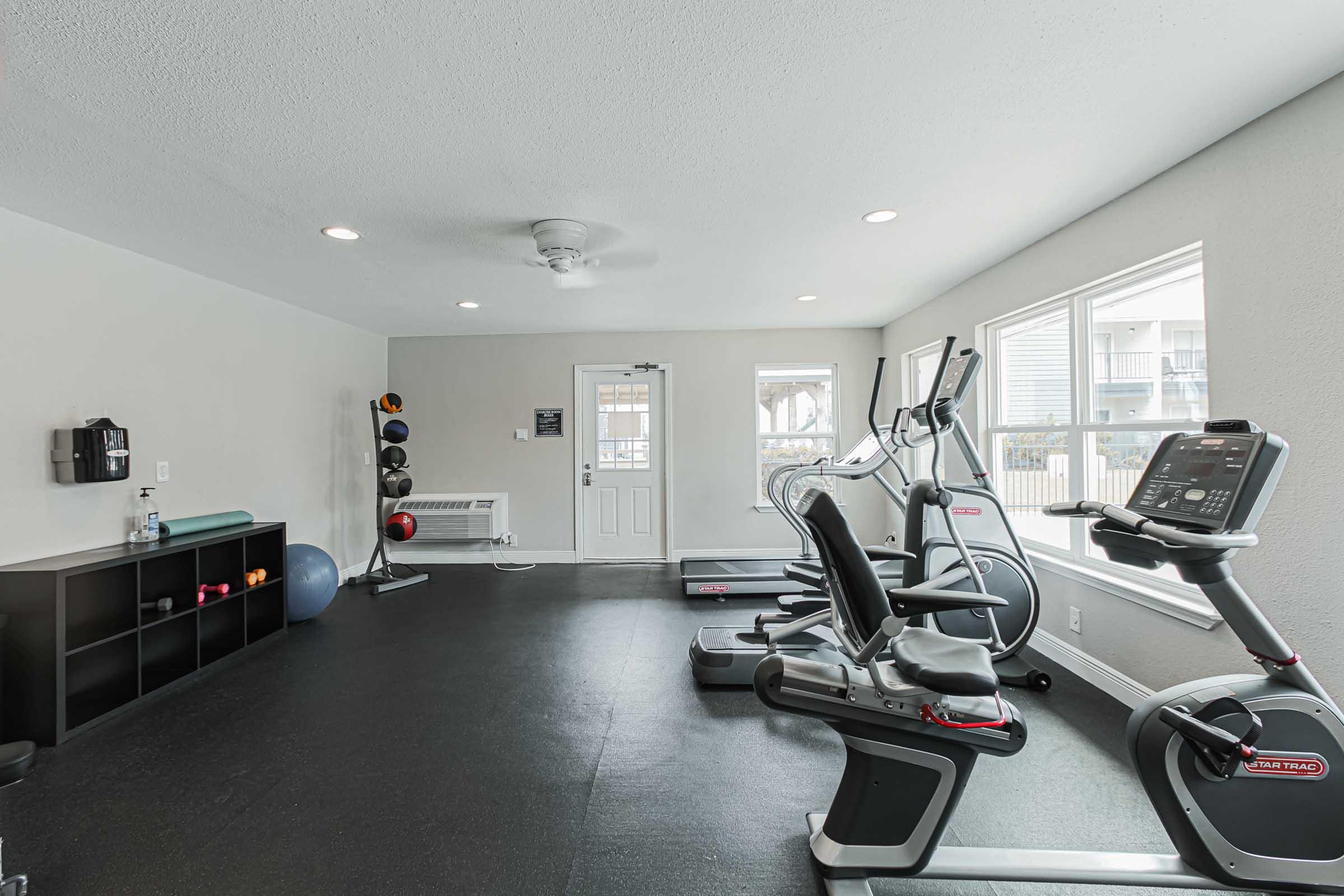 A bright and clean fitness room featuring various exercise equipment. There are two stationary bikes, an elliptical machine, and a treadmill, all arranged on a black floor mat. The walls are painted light gray, and large windows allow natural light to fill the space. A water bottle and a shelf with fitness accessories are visible.