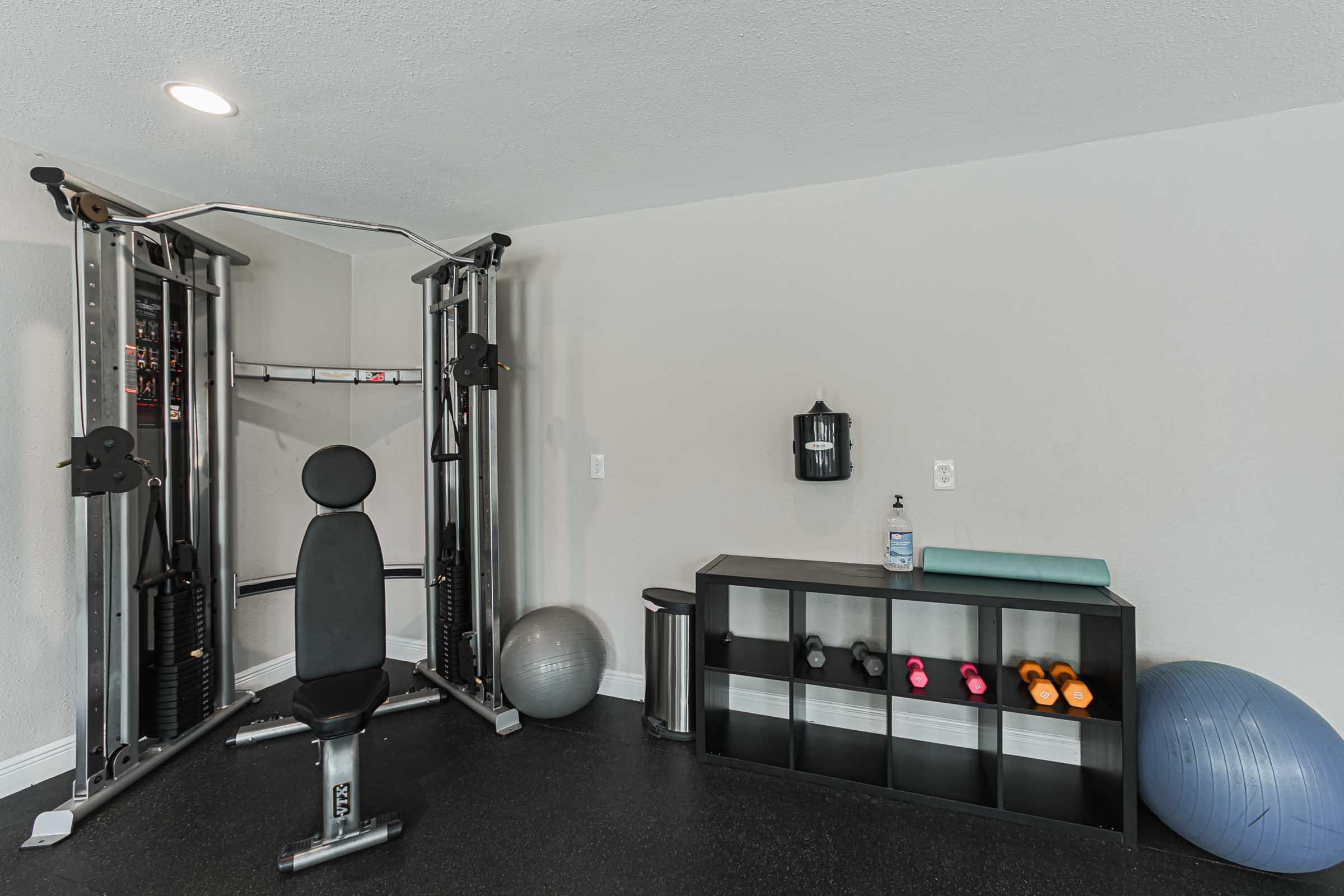 A home gym setup featuring a multi-station exercise machine, a black storage shelf with colorful dumbbells, a water bottle, a yoga mat, and stability balls. The space has a modern design with neutral-colored walls and flooring.