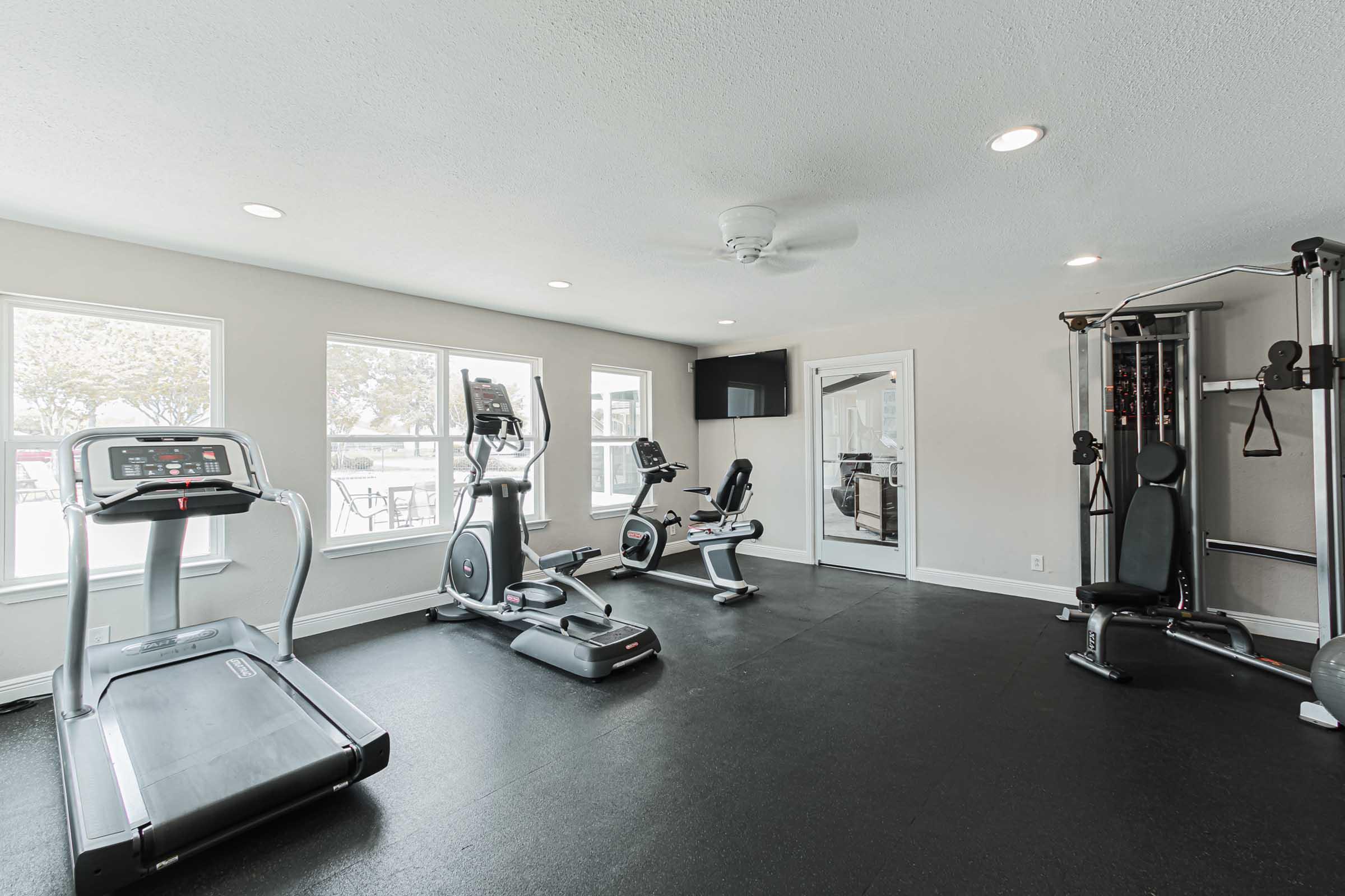 Interior view of a modern gym featuring various exercise equipment, including a treadmill, stationary bike, and a multi-gym machine. Bright natural light streams through several windows, illuminating the space, which has a neutral color palette and a ceiling fan for ventilation.