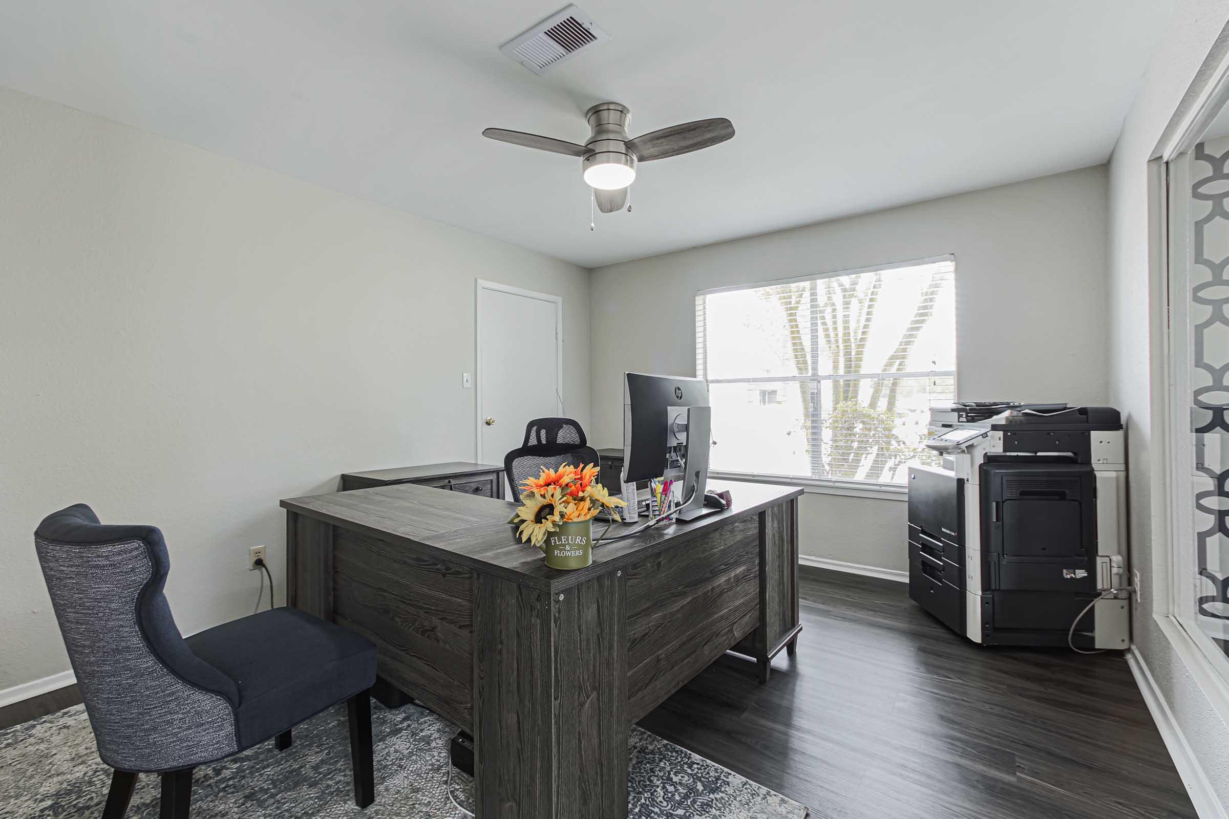 A modern office space featuring a wooden desk with a computer monitor, a comfortable chair, and a printer. The room has light-colored walls and a large window that lets in natural light, with a vase of orange flowers on the desk and a patterned window covering for added decor.
