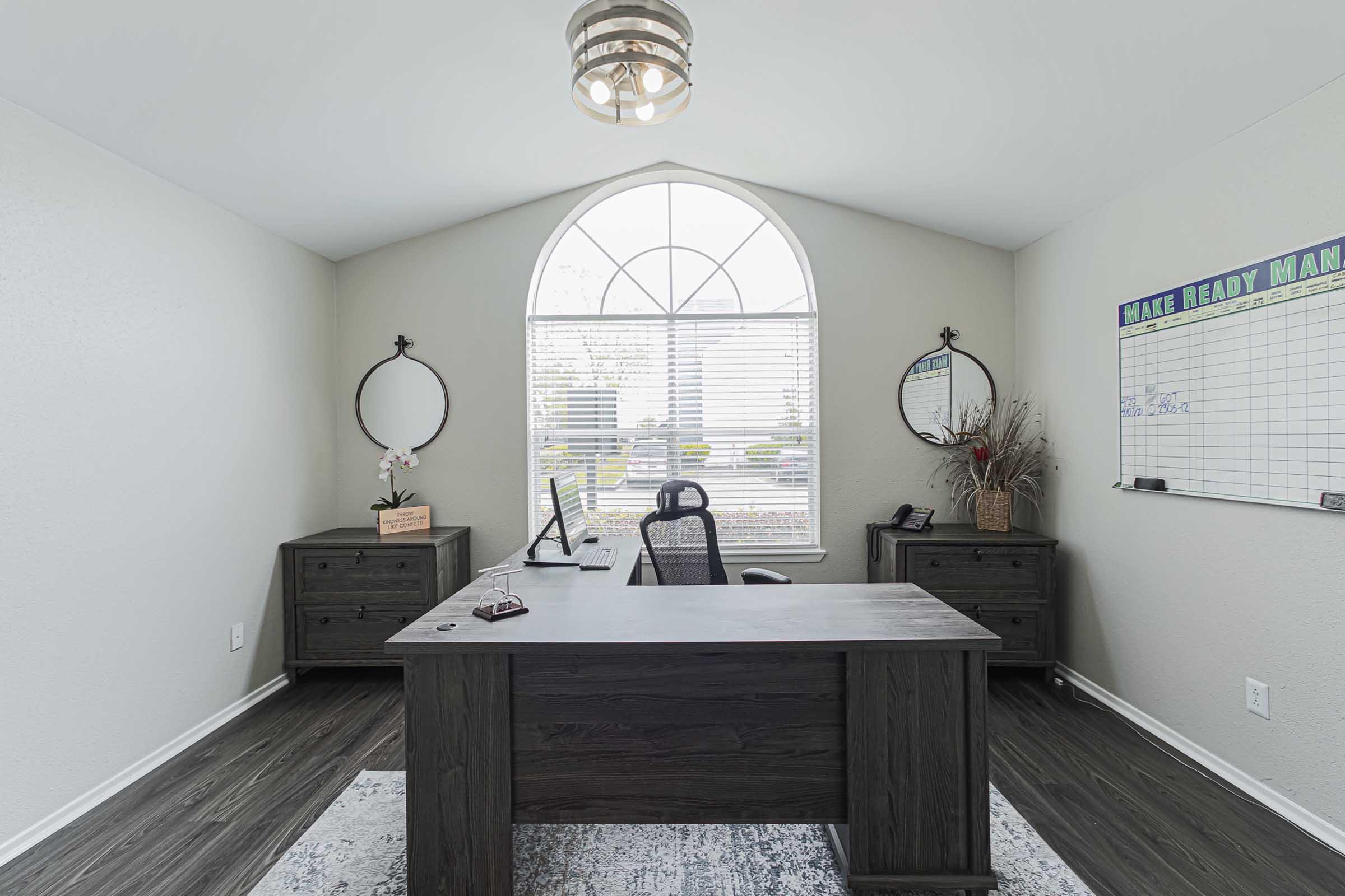 A modern office space featuring a dark wooden desk with a black office chair. Two round mirrors are mounted on the walls, and a whiteboard with a header is visible. Natural light floods the room through a large arched window, illuminating the minimalist decor and wooden flooring.
