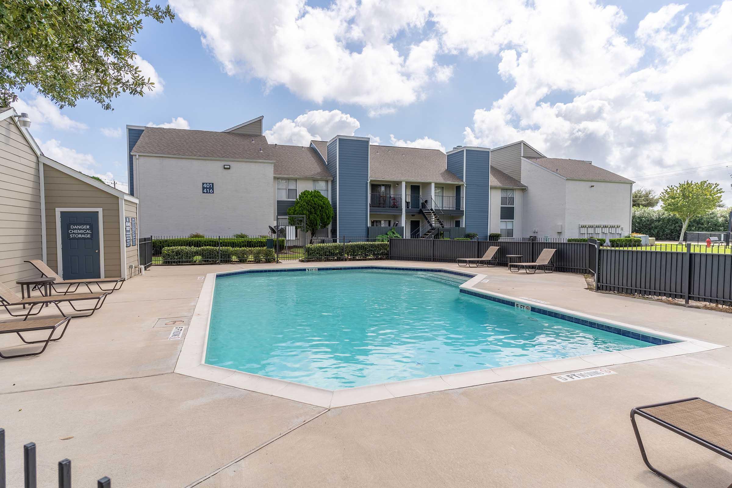 A swimming pool surrounded by lounge chairs, set in a residential area. The pool is clear and inviting, with blue skies and fluffy clouds overhead. Nearby, there are multi-story apartment buildings with green landscaping, creating a relaxed atmosphere.