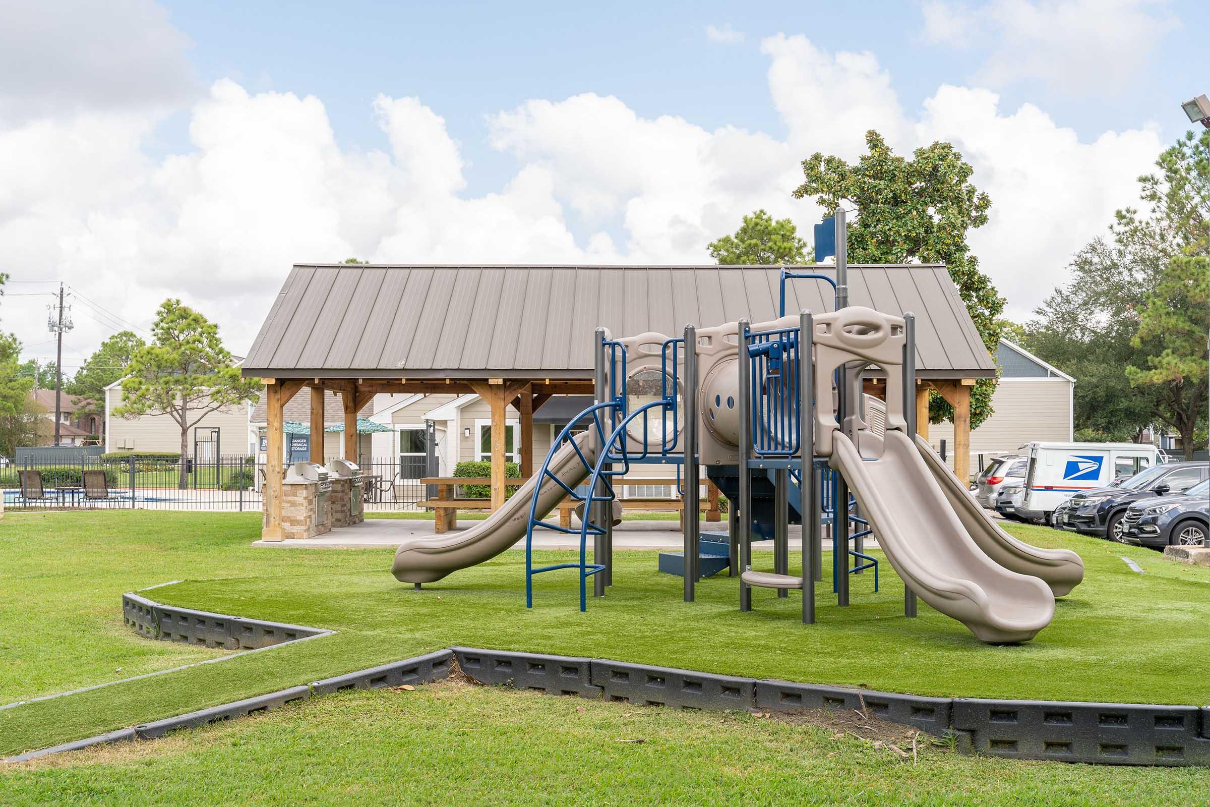 A playground featuring a slide and climbing structure, set on green synthetic grass. In the background, there is a covered picnic area and nearby vehicles parked. The sky is partly cloudy, creating a bright and inviting atmosphere for outdoor play.