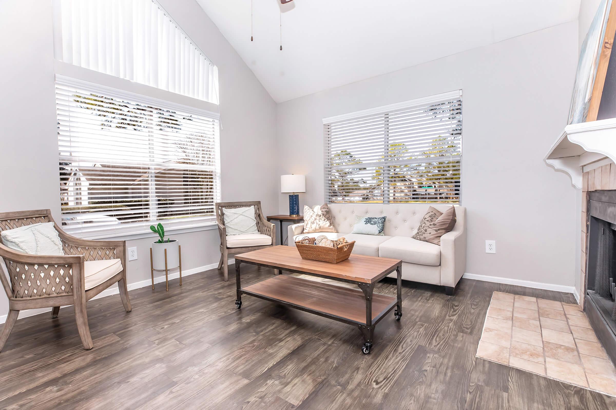 Bright and airy living room with large windows letting in natural light. Features a beige couch, a wooden coffee table with a basket, and two stylish chairs. A lamp provides additional lighting, and the floor has a mix of wood and tile. Modern decor enhances the inviting atmosphere.