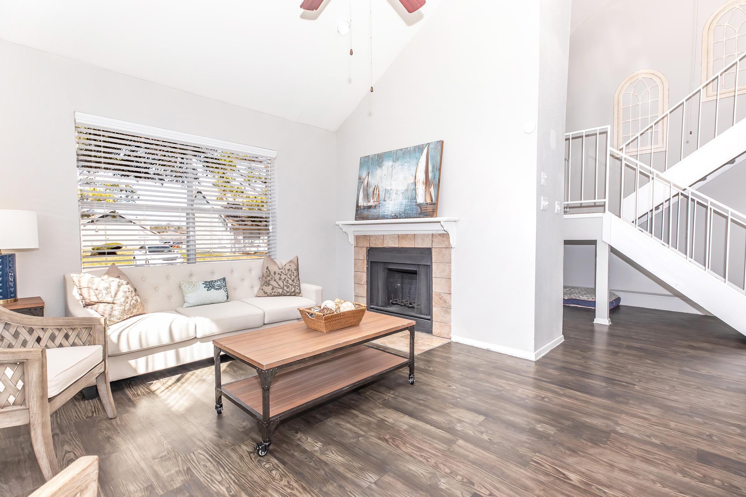 A bright and airy living room featuring a light-colored sofa with decorative pillows, a wooden coffee table with a basket, and a cozy fireplace. Large windows with blinds allow natural light in, while a staircase leads to an upper level. The flooring is dark wood, adding warmth to the space.
